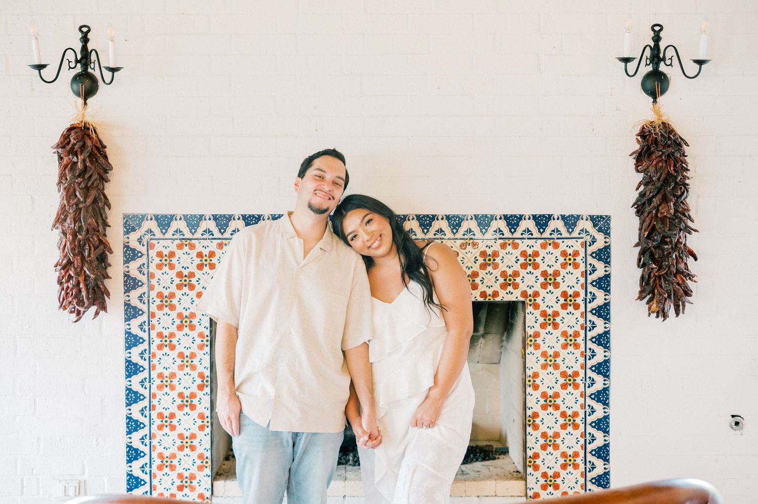 A couple poses together in front of a white wall decorated with ornate tiles and hanging dried peppers.