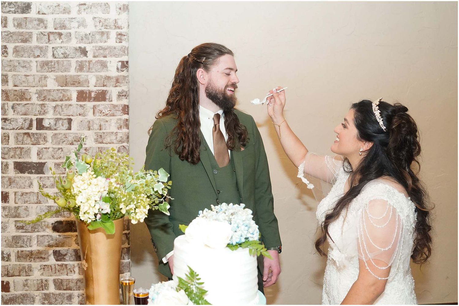 Bride feeds groom wedding cake at reception, white tiered cake displayed on table with floral arrangement, brick wall backdrop.
