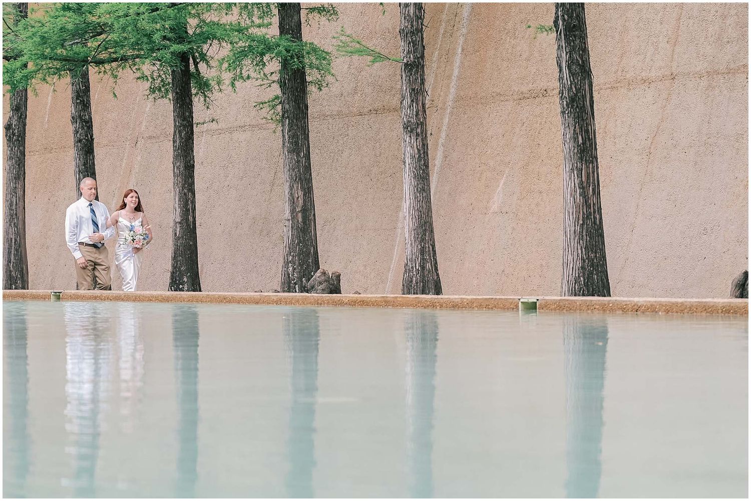 Tree-lined pathway with beige stone wall and ground creating a serene outdoor corridor.