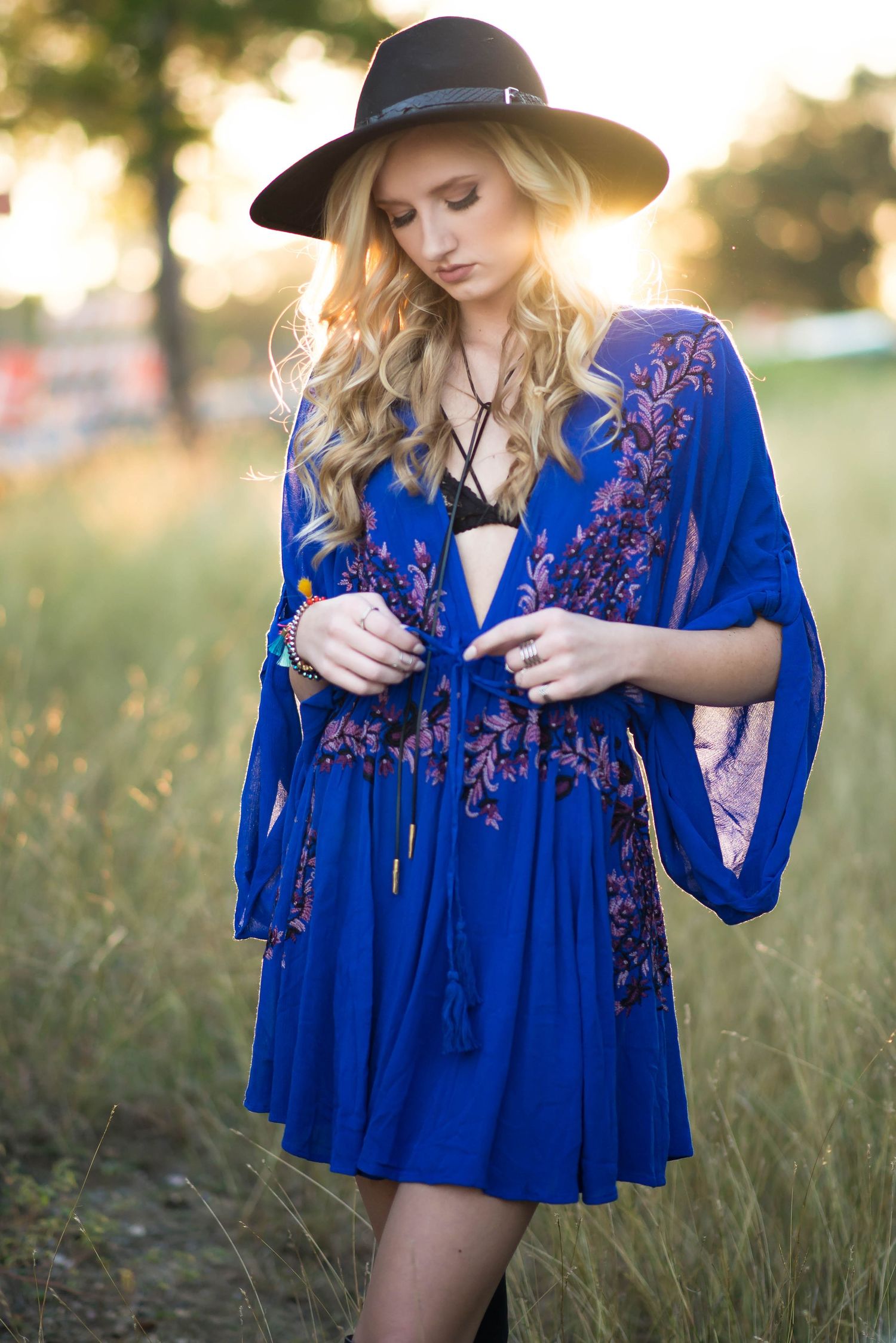 A bohemian style royal blue dress with floral embroidery and bell sleeves is worn with a black wide-brimmed hat in a field. Seymour TX HS