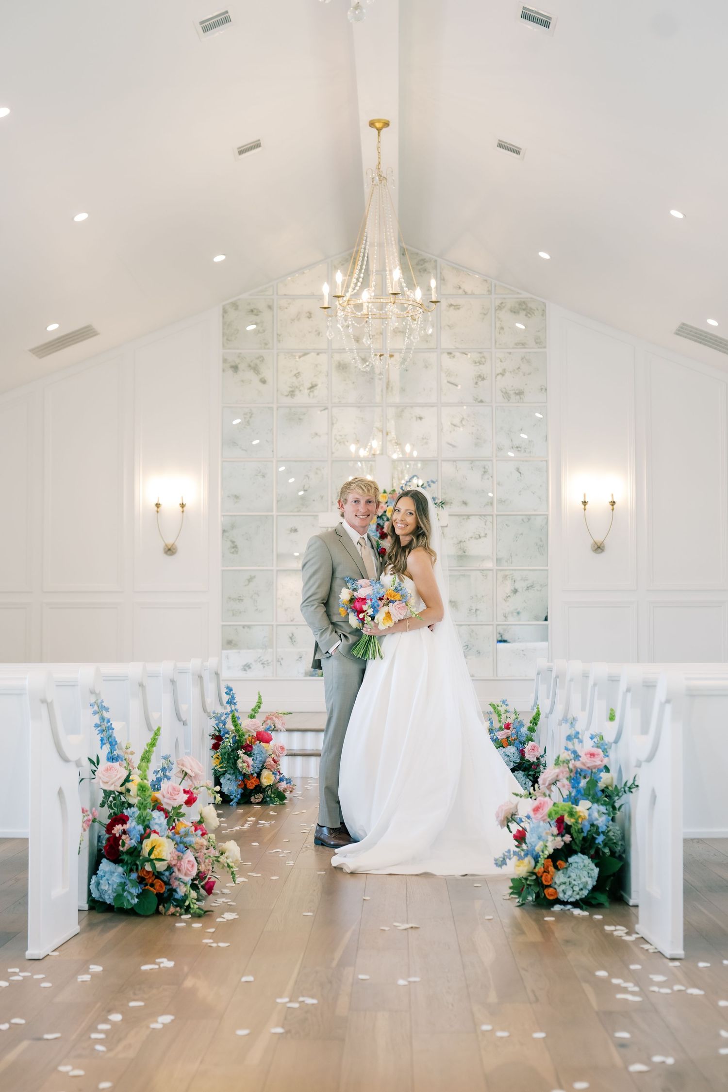 Bride and groom pose in elegant white chapel with colorful floral arrangements lining the aisle and a crystal chandelier above.