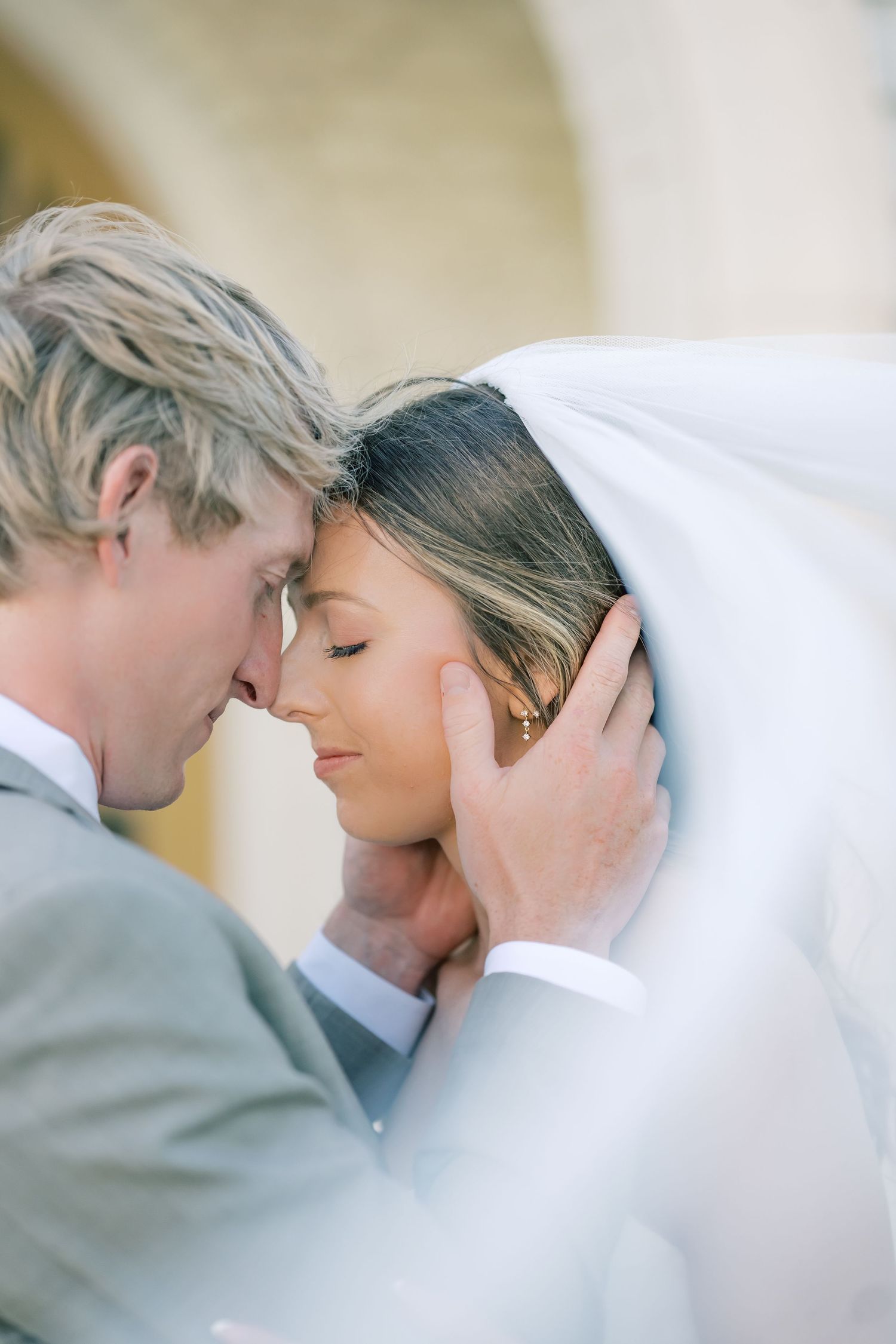 A groom in a gray suit tenderly holds his bride's face under her veil, foreheads touching in an intimate wedding moment.