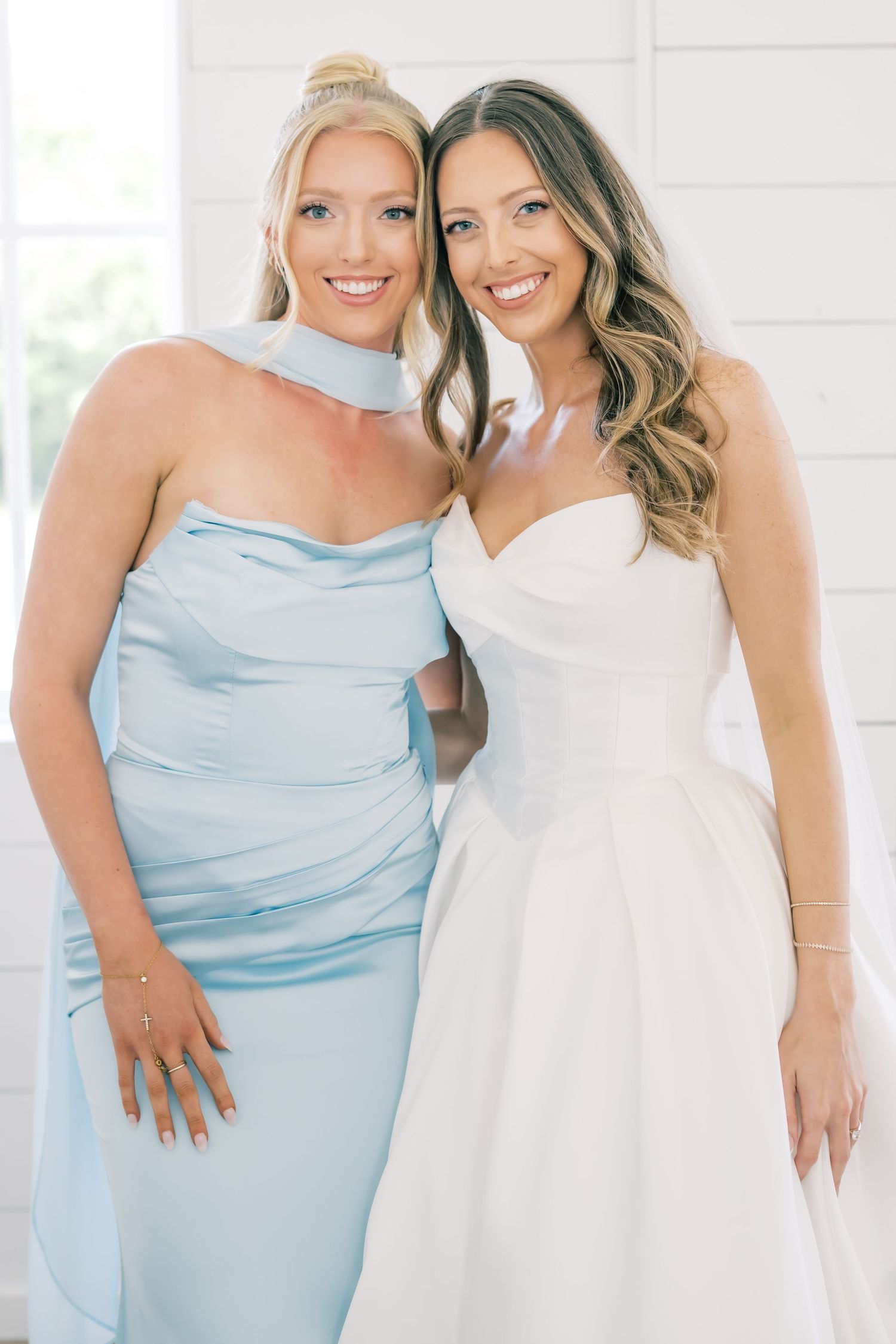 Bride in strapless white gown poses with bridesmaid in light blue satin dress indoors.