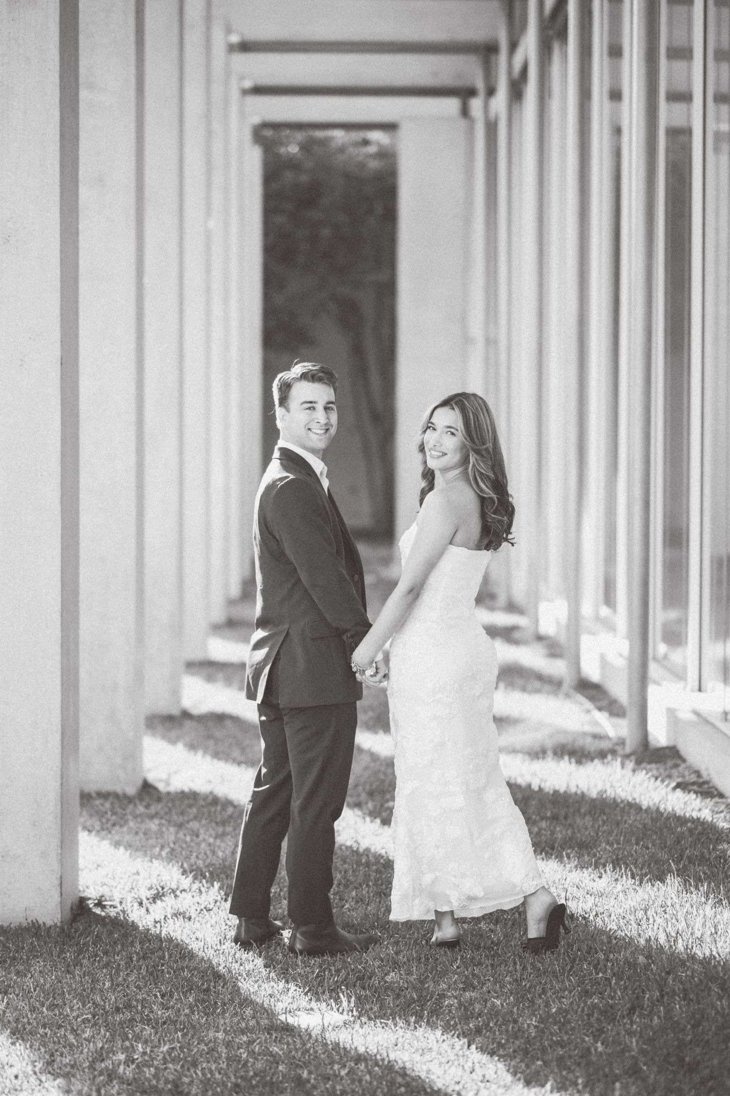 A bride and groom share a playful moment walking between white pillars in black and white photography.
