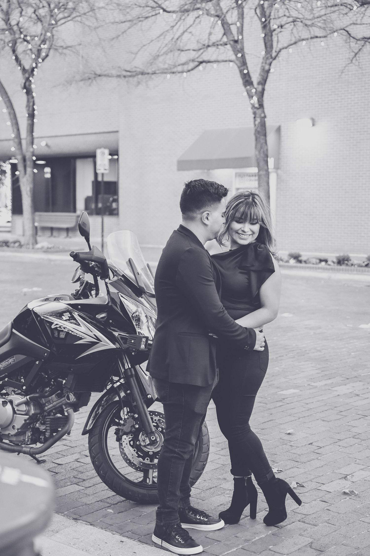 Black and white photo of a couple embracing and laughing together in front of a motorcycle on a brick street.