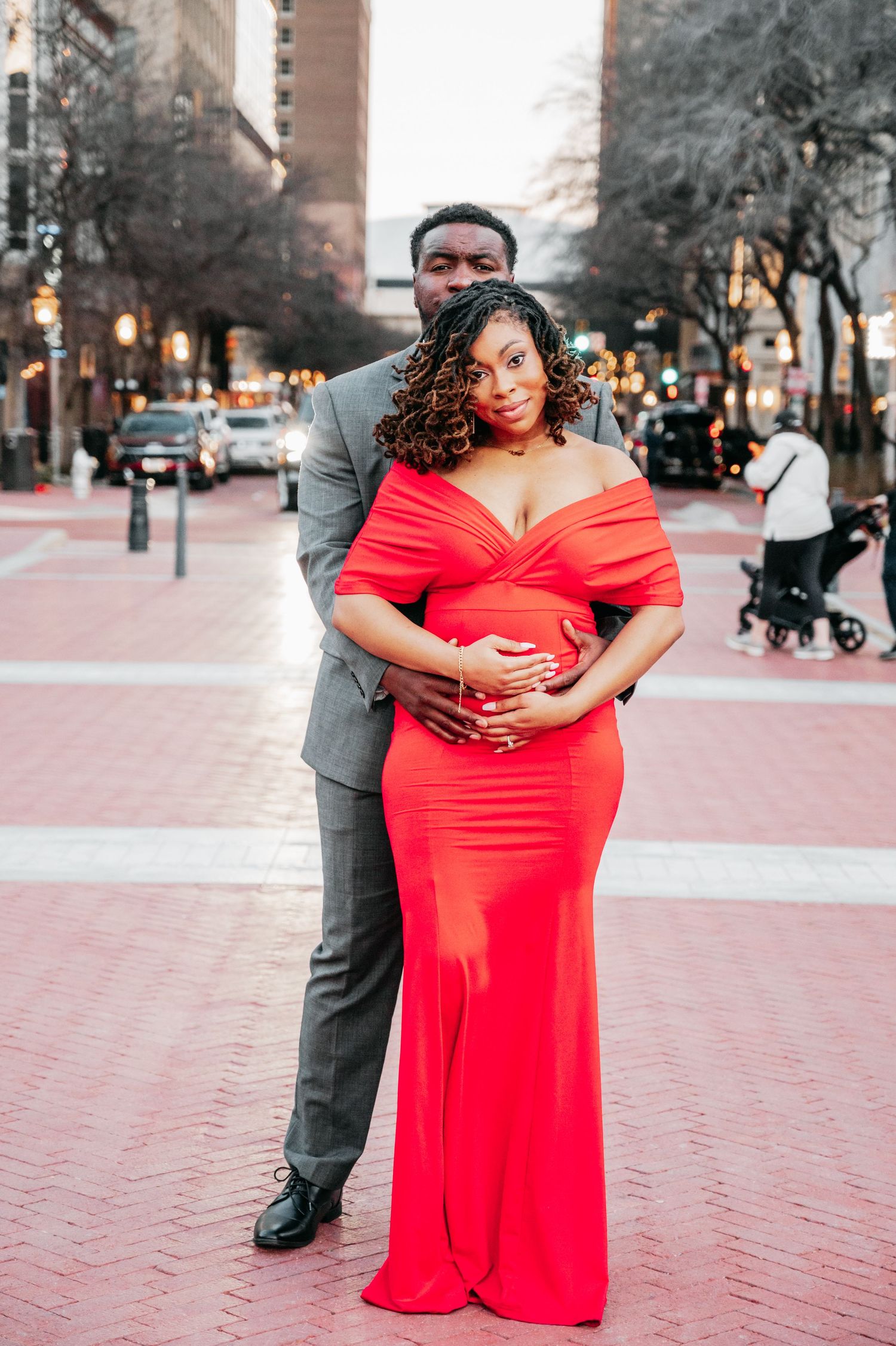 Couple poses on urban brick plaza, woman in red off-shoulder maternity gown, man in gray suit embracing her from behind.