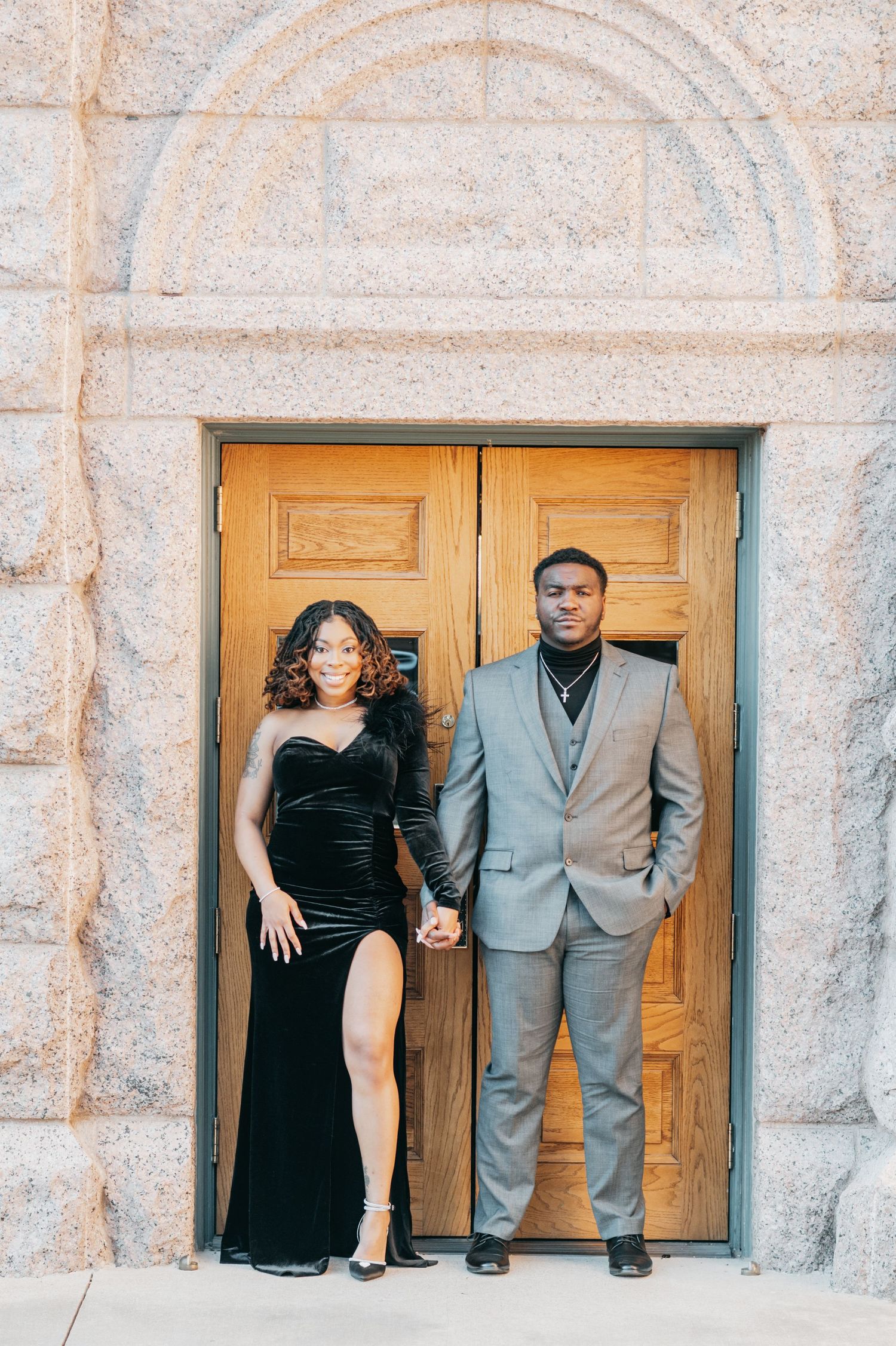 Couple posing hand-in-hand before ornate wooden doors, woman in black velvet gown and man in gray suit.