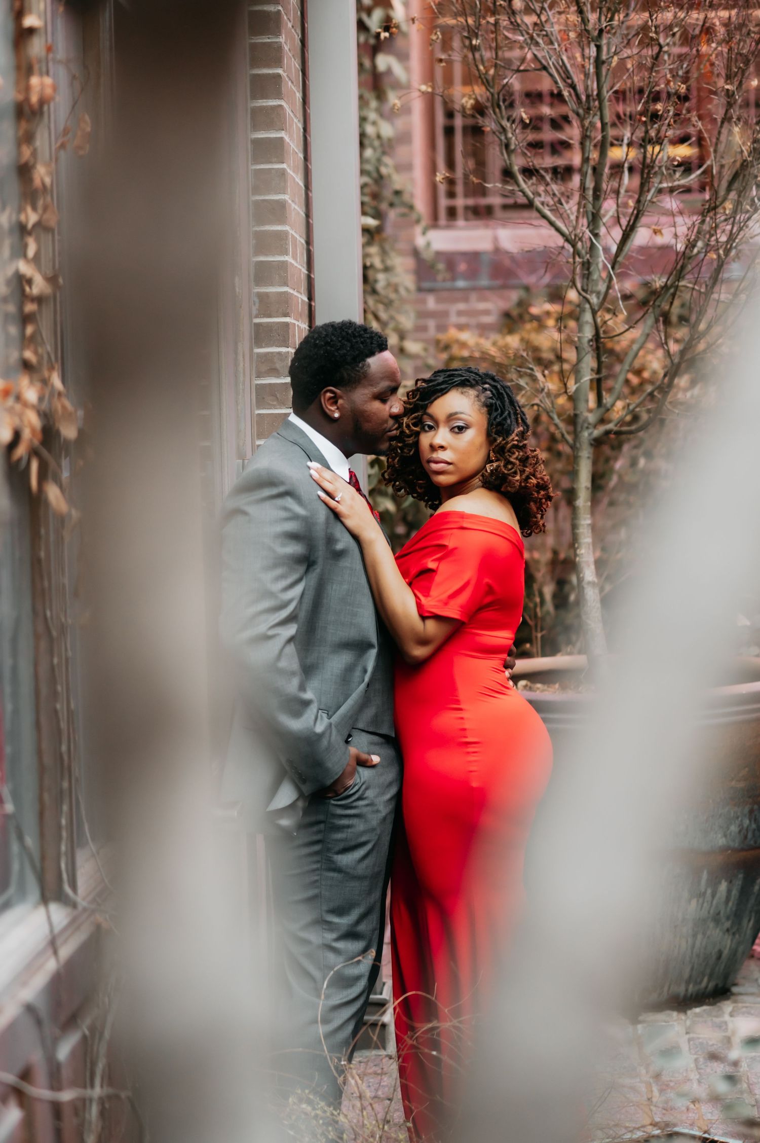 A couple in elegant attire, man in gray suit and woman in red dress, share a romantic embrace outdoors.