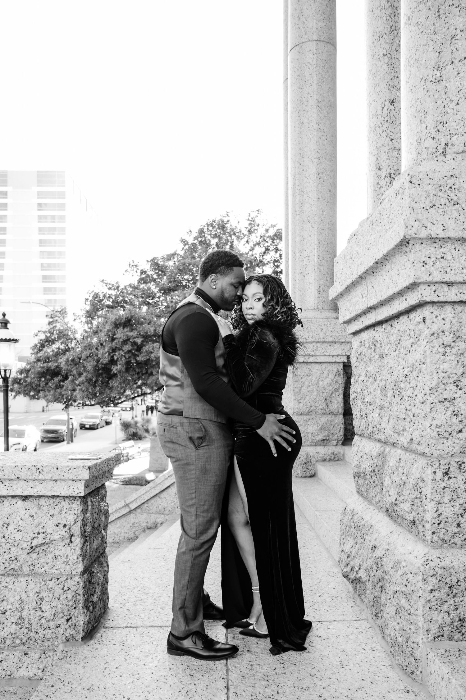 Black and white photo of a couple in elegant attire sharing a romantic moment beside stone columns outdoors.