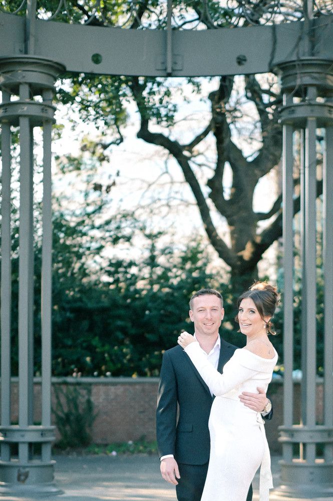 Couple poses under ornate metal gazebo, bride in white off-shoulder gown, groom in black suit, lush greenery background.