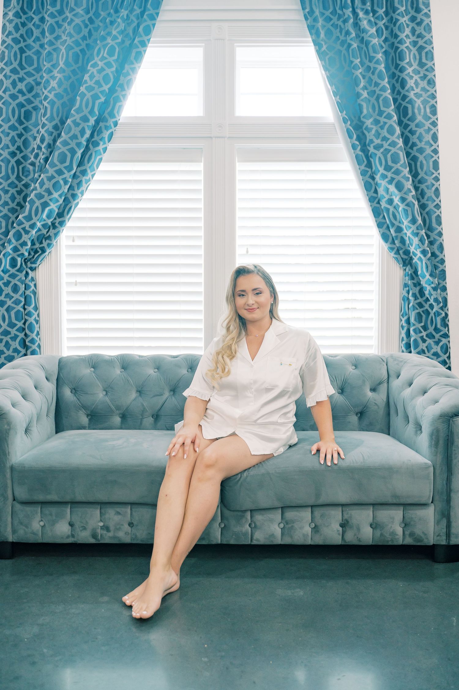 Woman in white robe sitting on tufted blue velvet sofa near window with teal curtains.