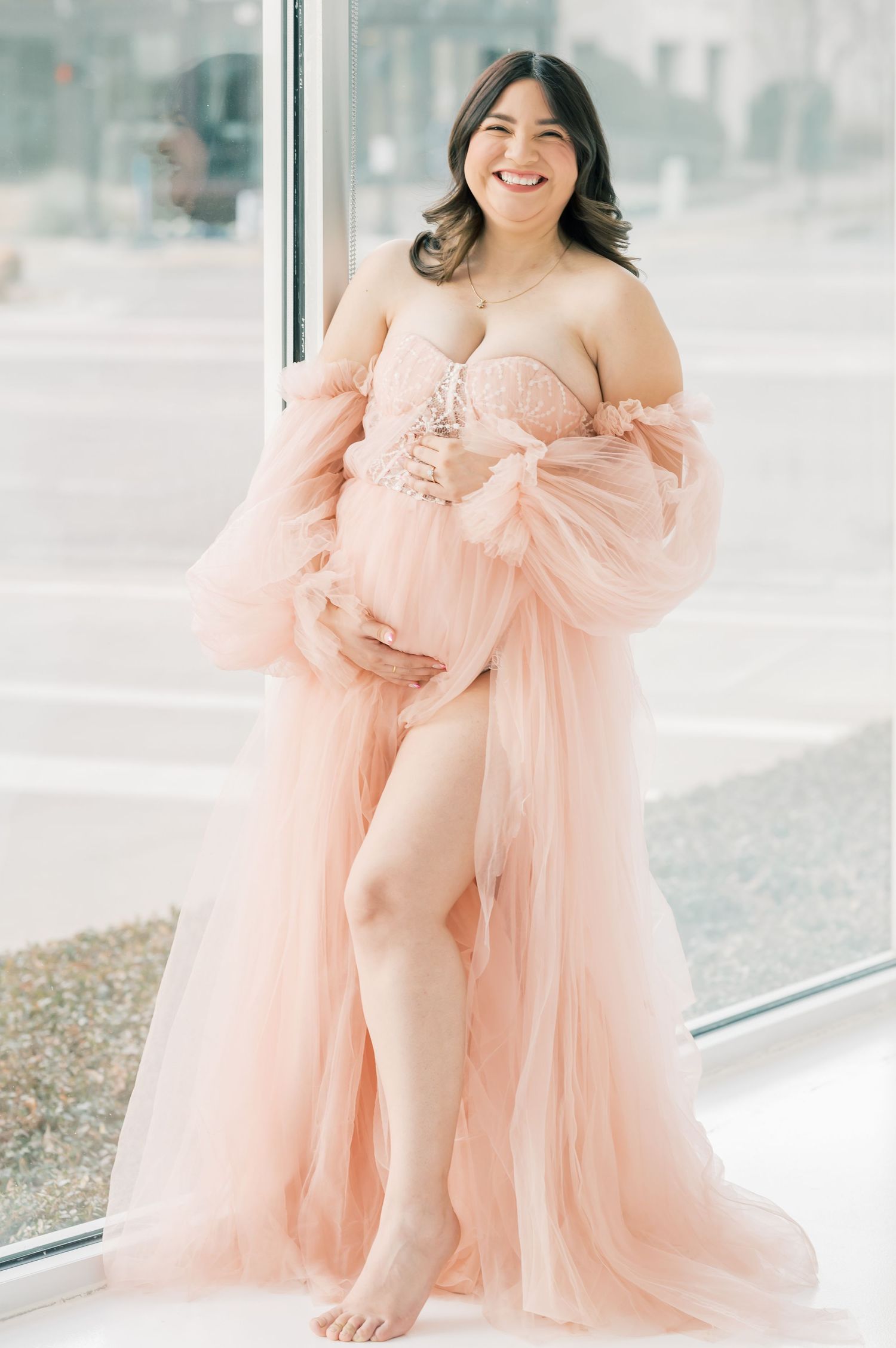Smiling pregnant woman wearing a blush pink off-shoulder tulle maternity gown poses by a window.