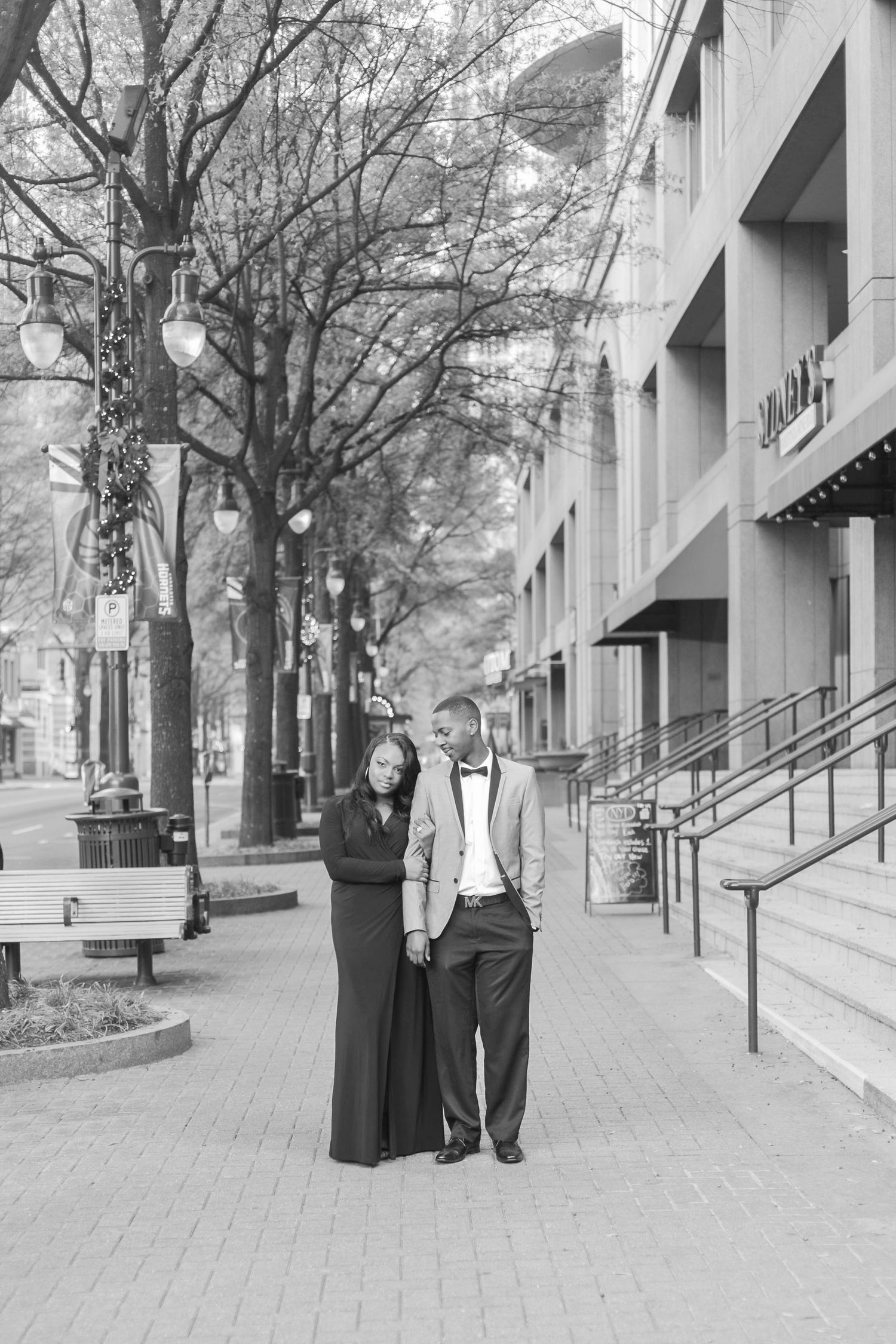 Black and white photo of couple in formal attire walking arm in arm on a city sidewalk.