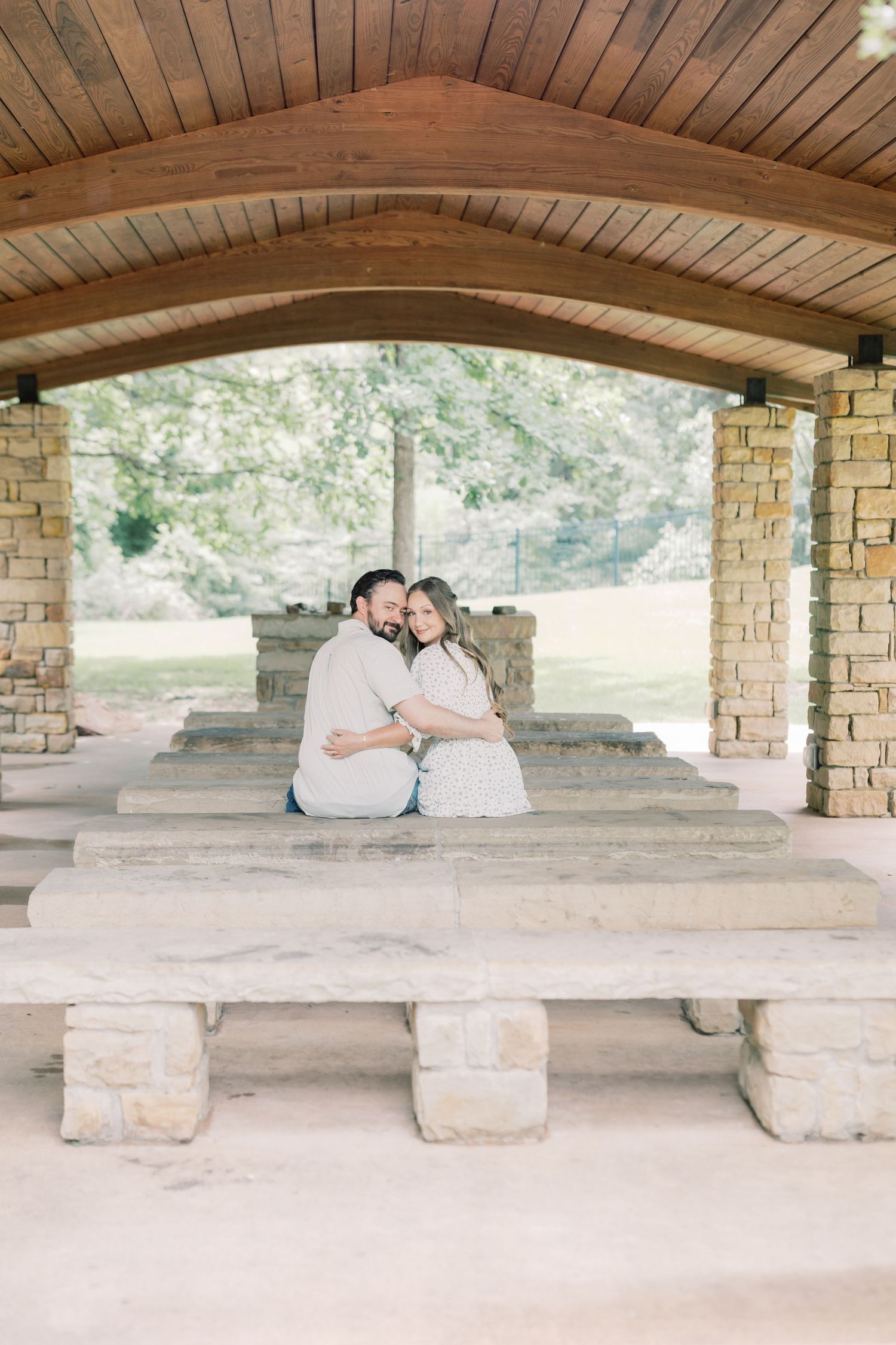 Couple sits closely together under a rustic stone and wood pavilion, sharing a tender embrace.