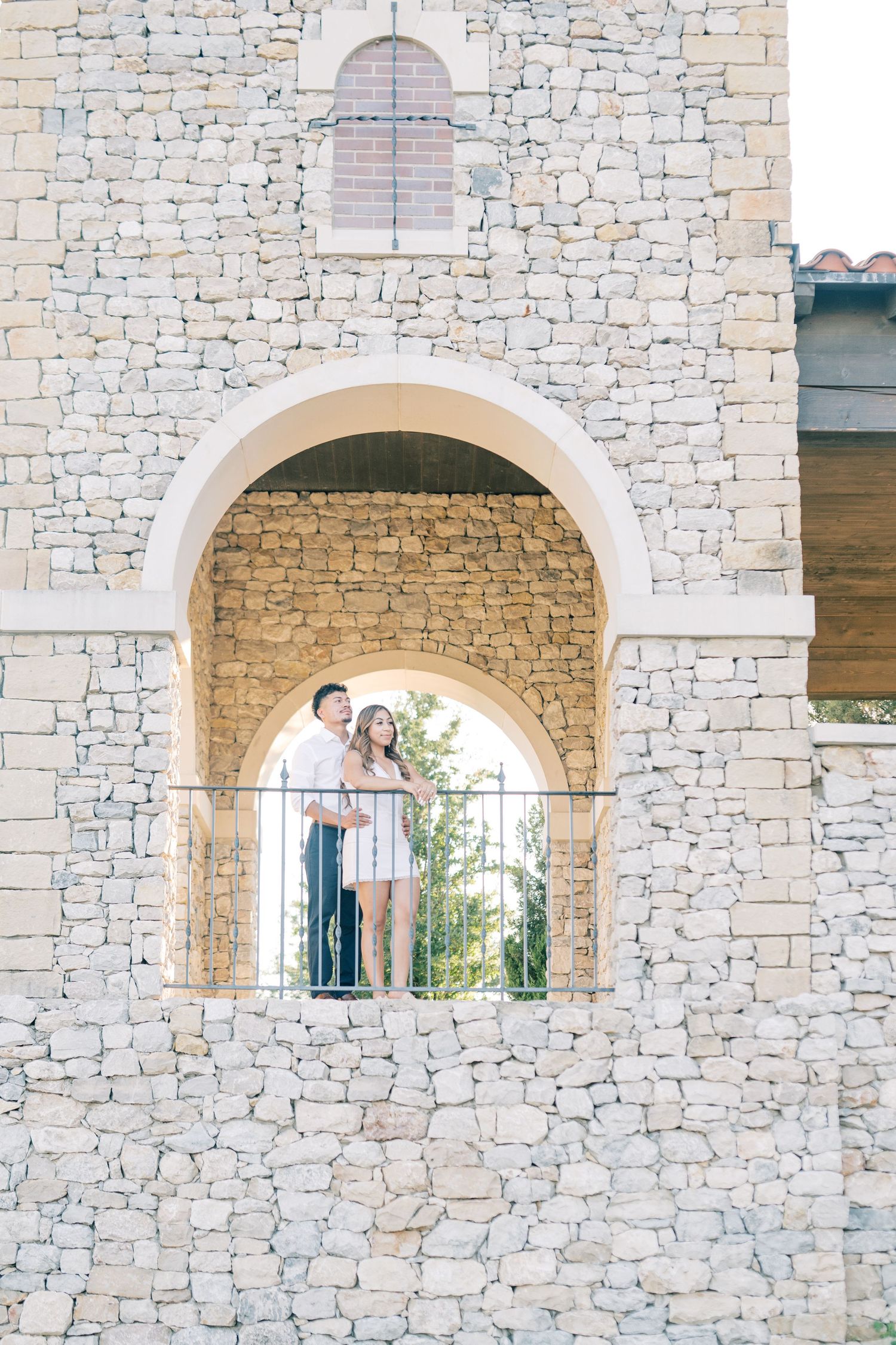 Romantic couple standing together in an arched stone balcony overlooking lush greenery.