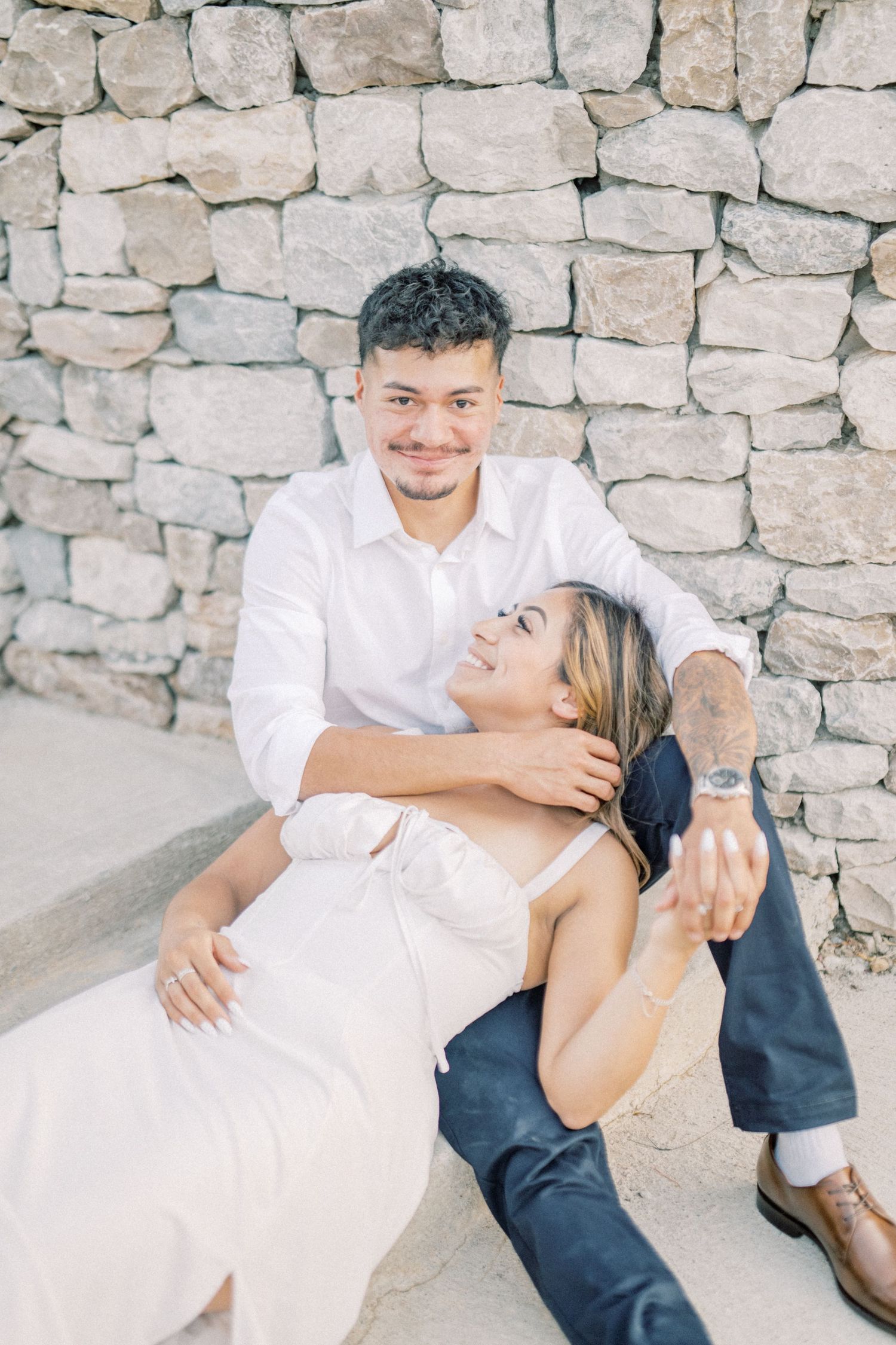 Smiling couple posing against a stone wall, woman reclining in mans arms during engagement photos.