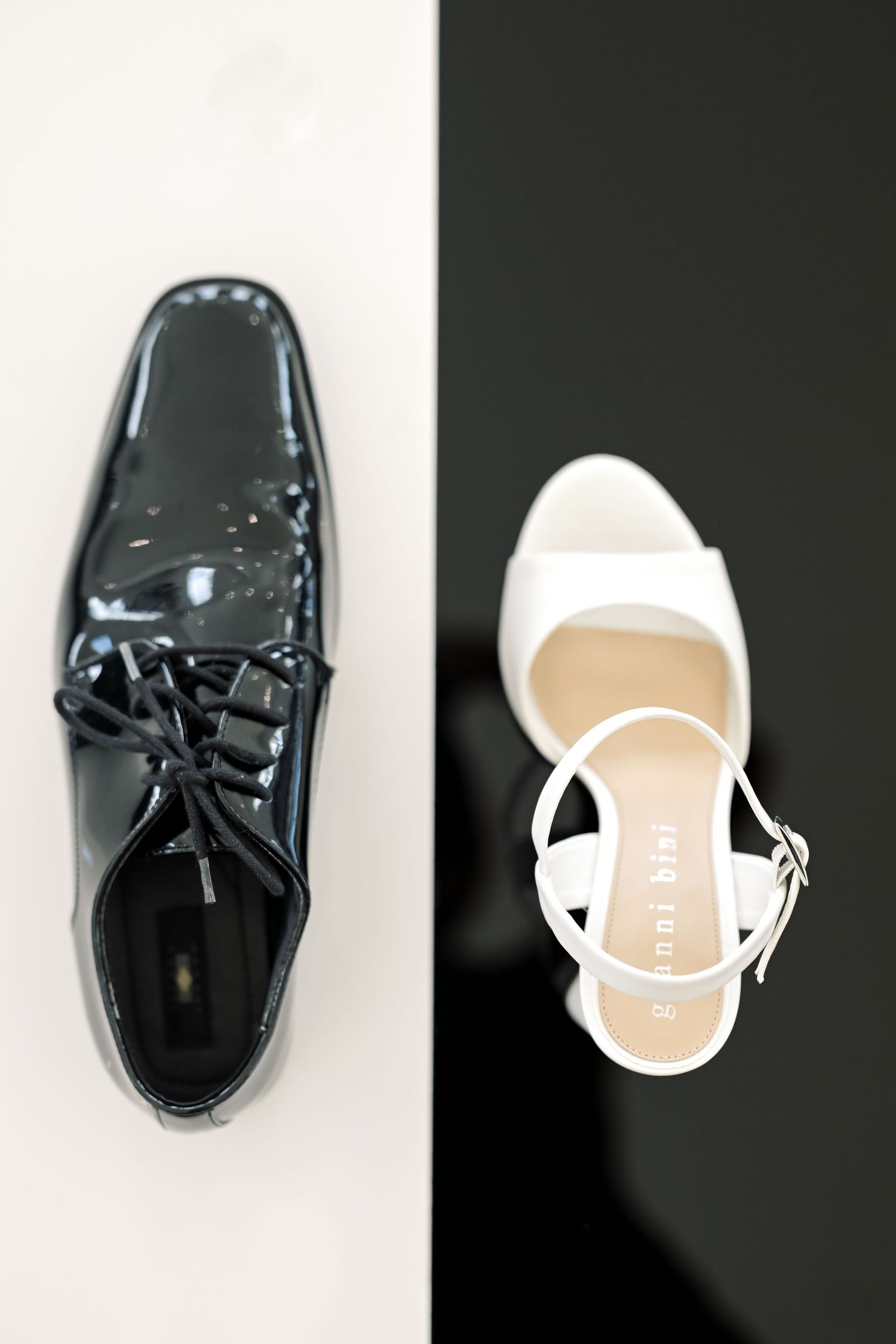 Black patent leather dress shoe beside a white strappy heeled sandal on contrasting black and white background.