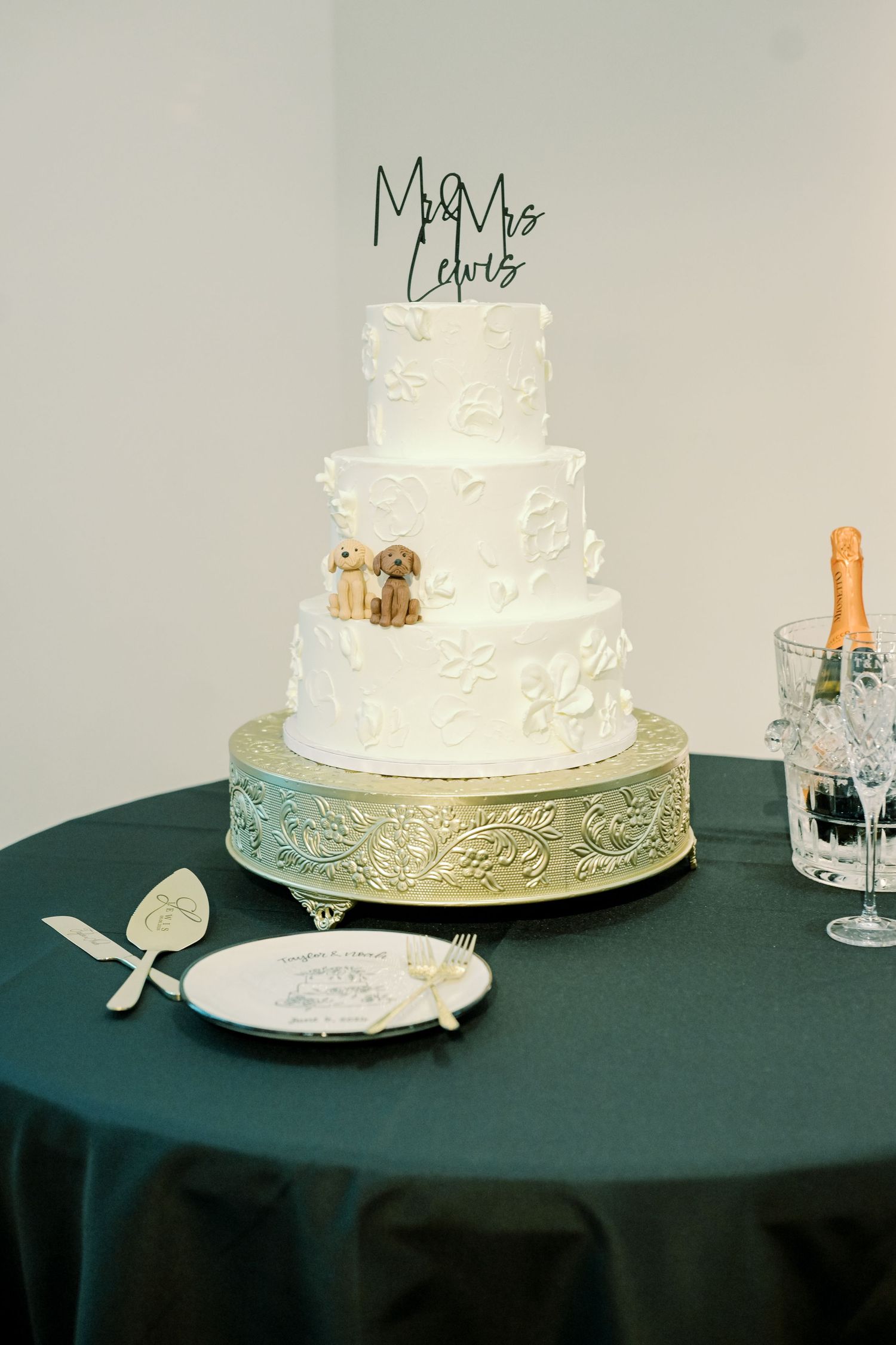 Three-tier white wedding cake with dog toppers and Mr & Mrs Lewis cake topper on gold stand.