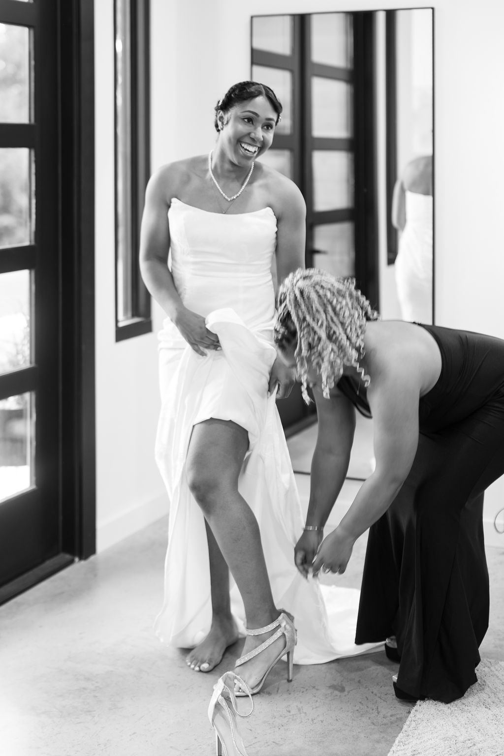 Black and white photo of a smiling bride in a white gown having her heels fastened by a bridesmaid.