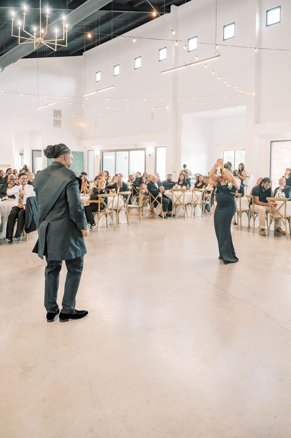 Couple shares first dance in bright modern wedding reception venue with string lights and seated guests.