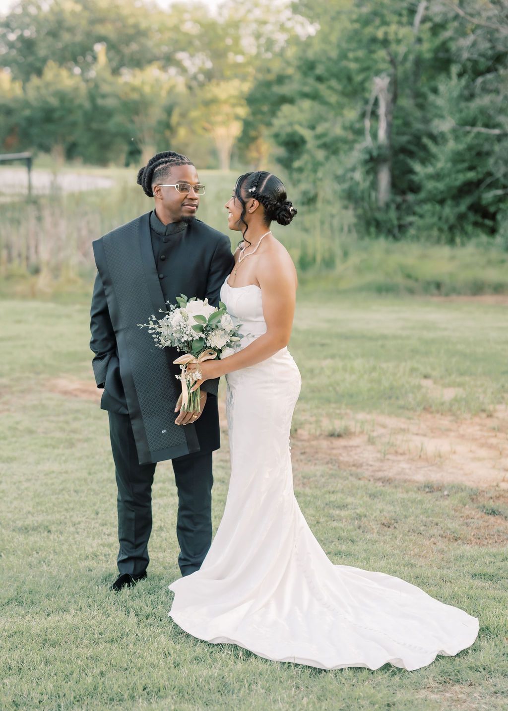 A bride and groom stand together on green grass, she in a white gown holding a bouquet, he in a black suit.