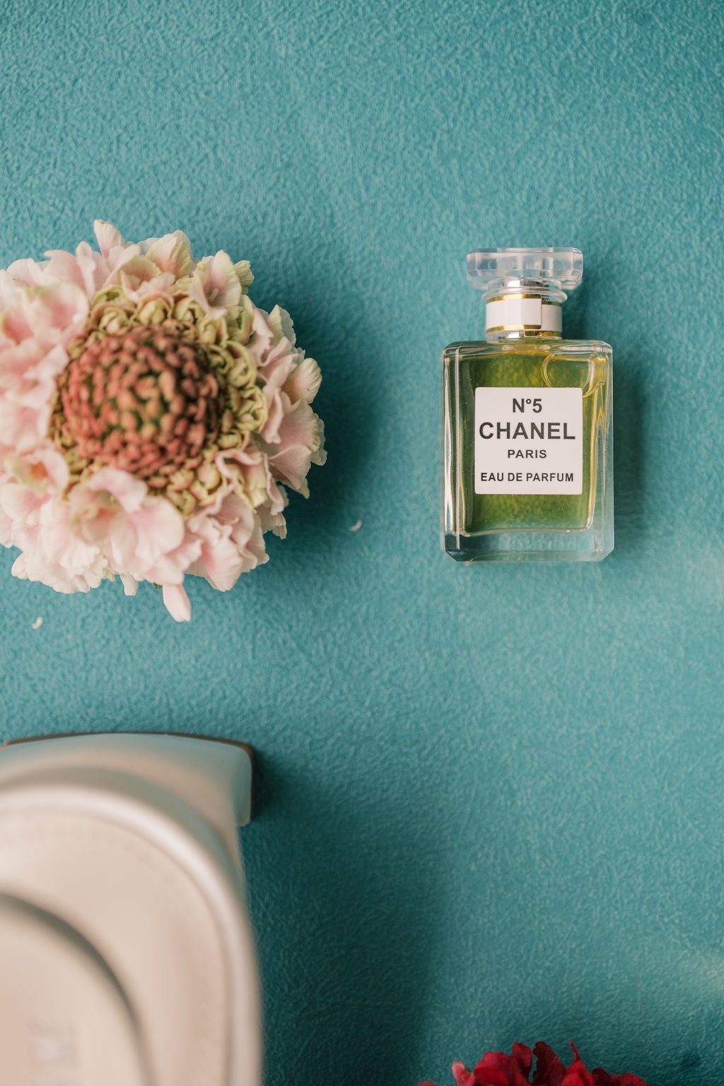 Chanel No. 5 Eau de Parfum bottle on teal wall beside pink flowers.