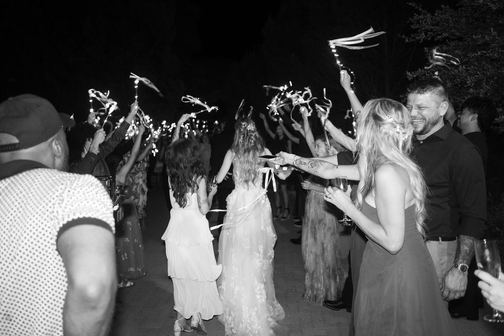Black and white photo of bride and groom walking through sparkler exit surrounded by cheering wedding guests at night.