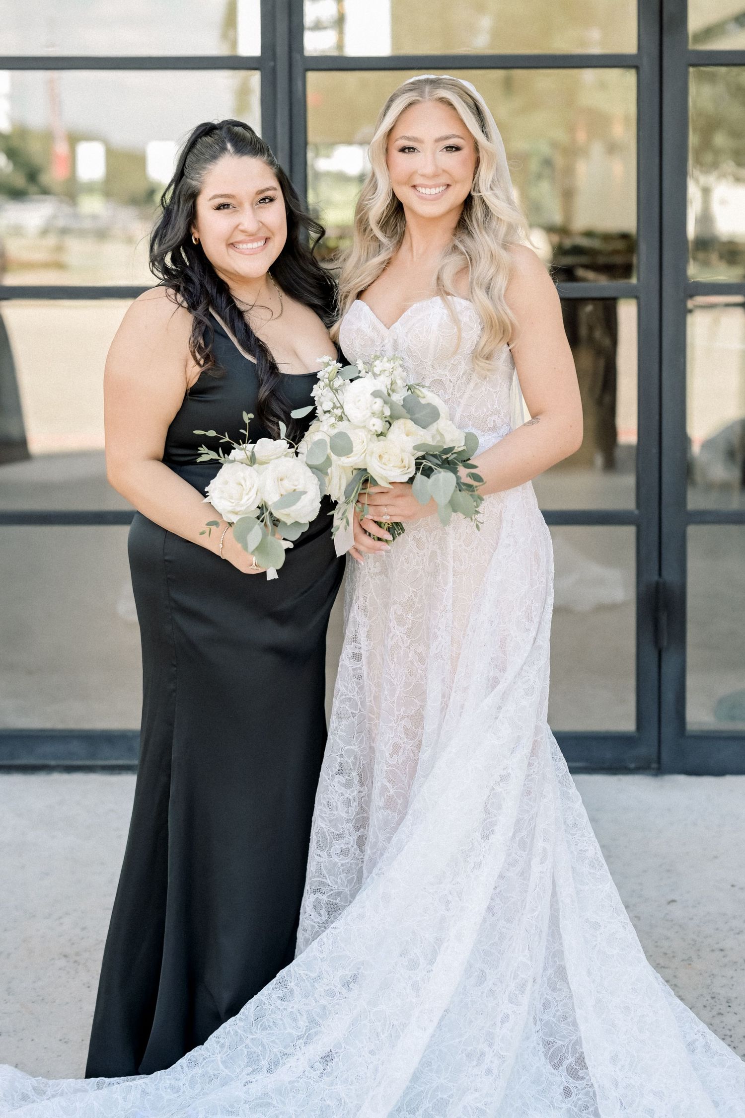 Bridesmaid in black satin dress stands with blonde bride holding white rose bouquet.