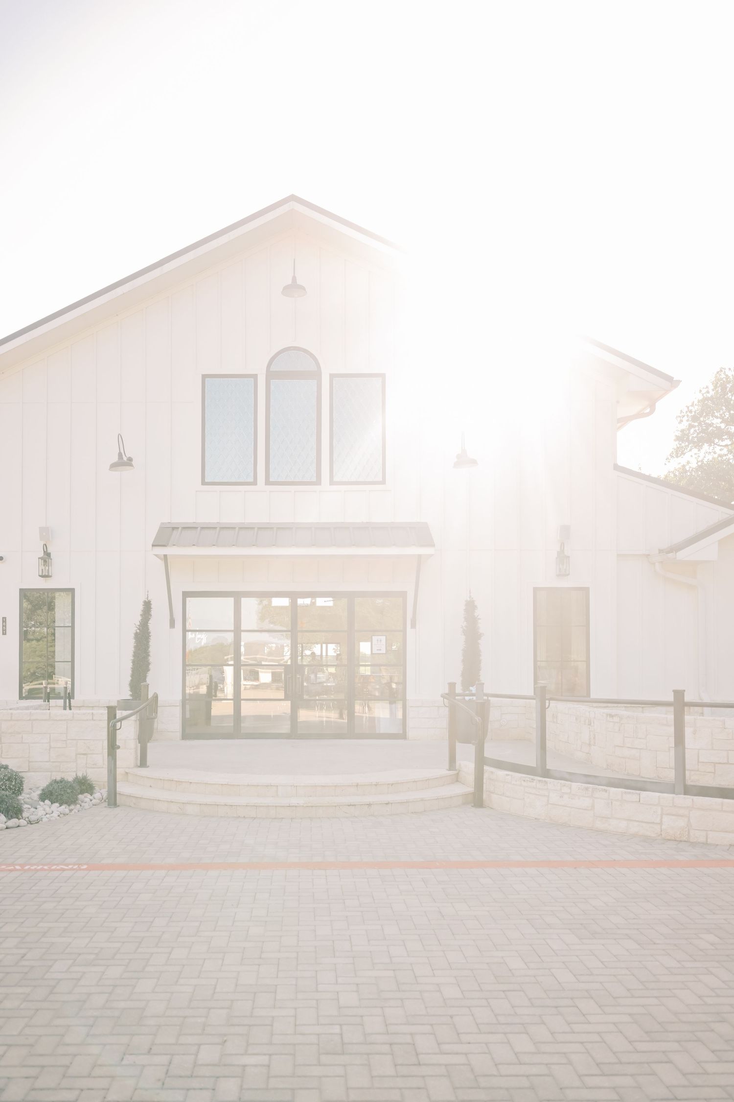 Bright sunlit exterior of white modern farmhouse wedding venue with arched windows and brick path.