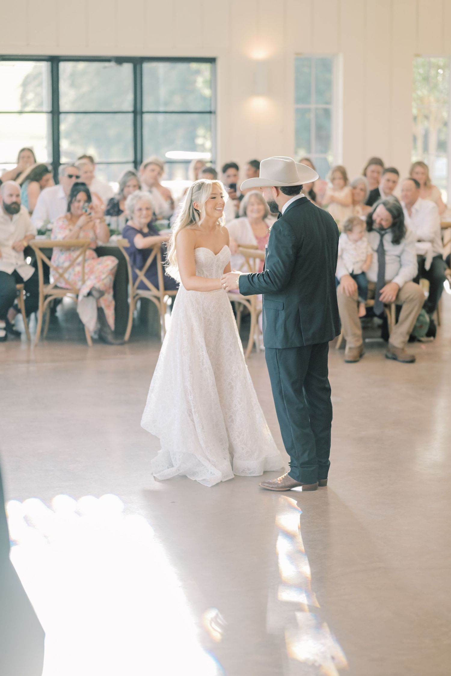 Bride and groom share first dance in bright reception hall before seated guests.