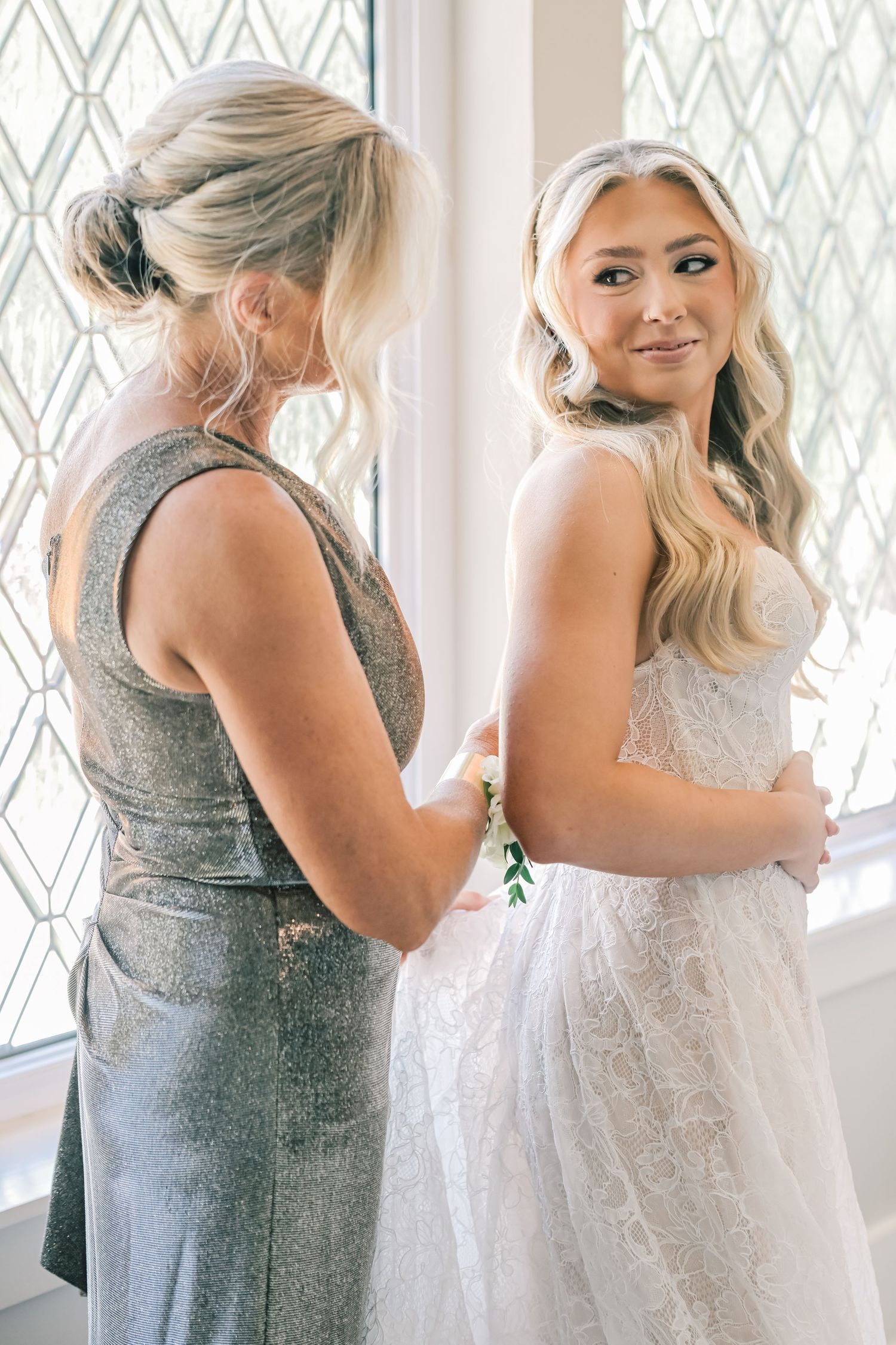 Mother helping bride into lace wedding gown before ceremony near leaded glass window.
