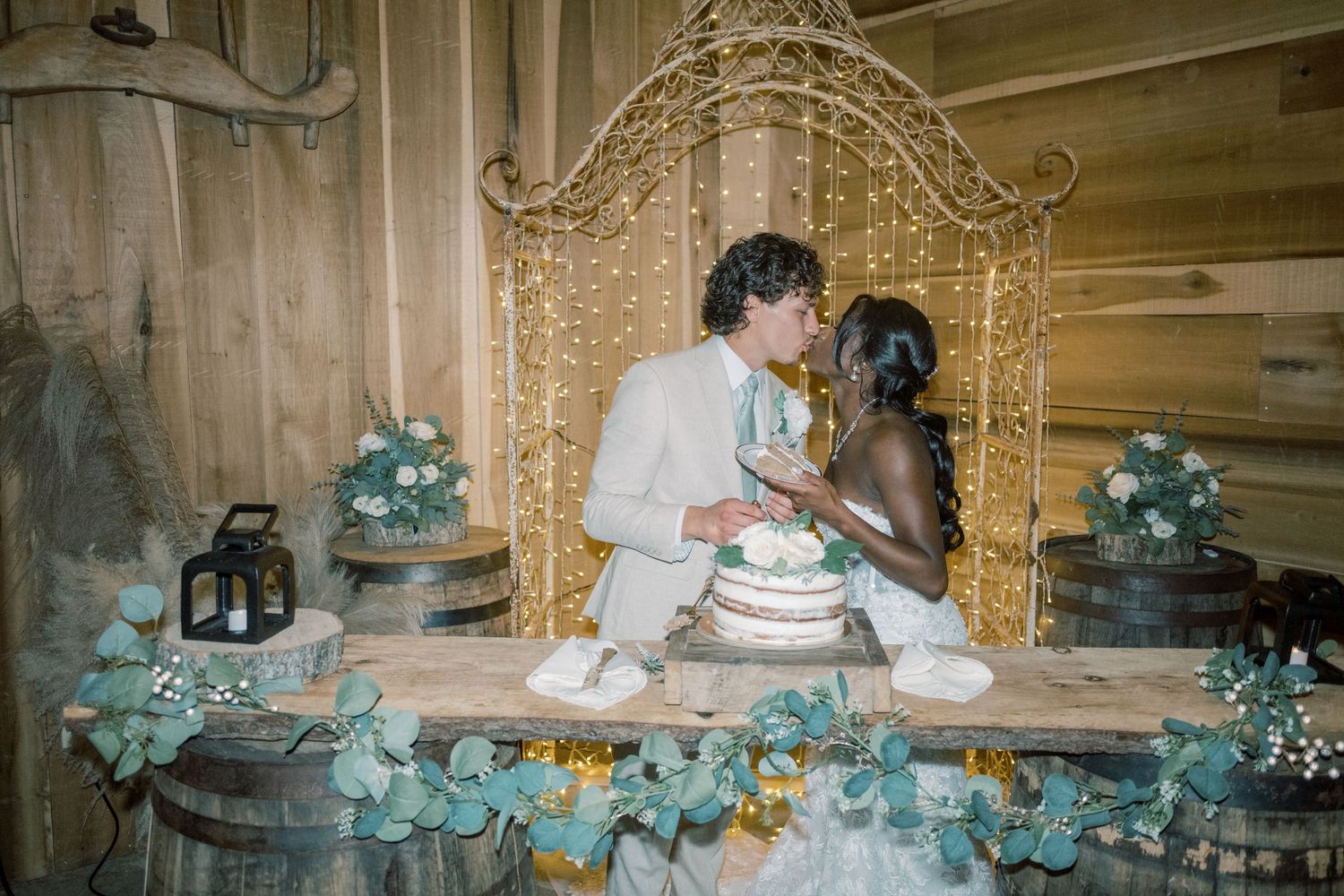 Newlyweds kiss while cutting naked wedding cake at rustic barn reception with fairy lights.
