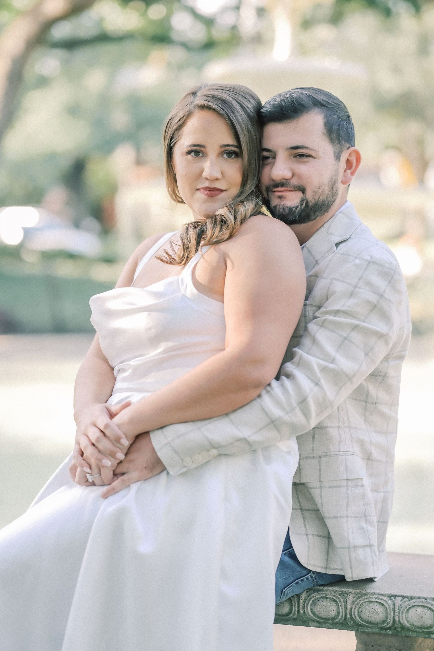 Couple in an engagement photo session, the man embracing the woman in a white dress from behind on a stone bench in a park.