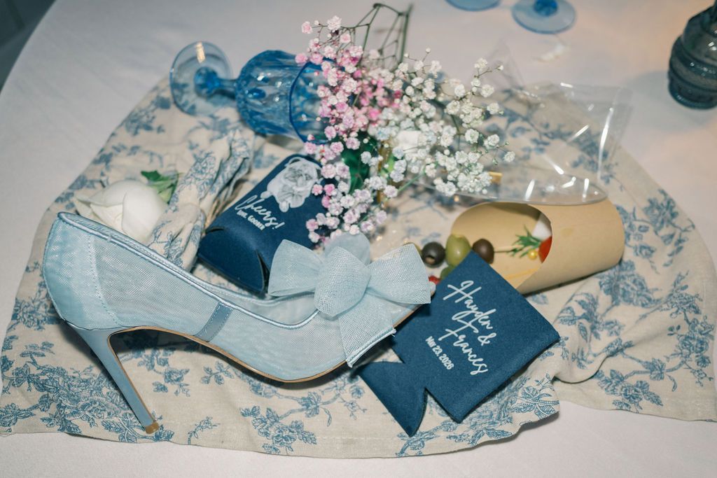 Blue lace wedding heels with bow displayed alongside floral bouquet and personalized gift tags on toile fabric.