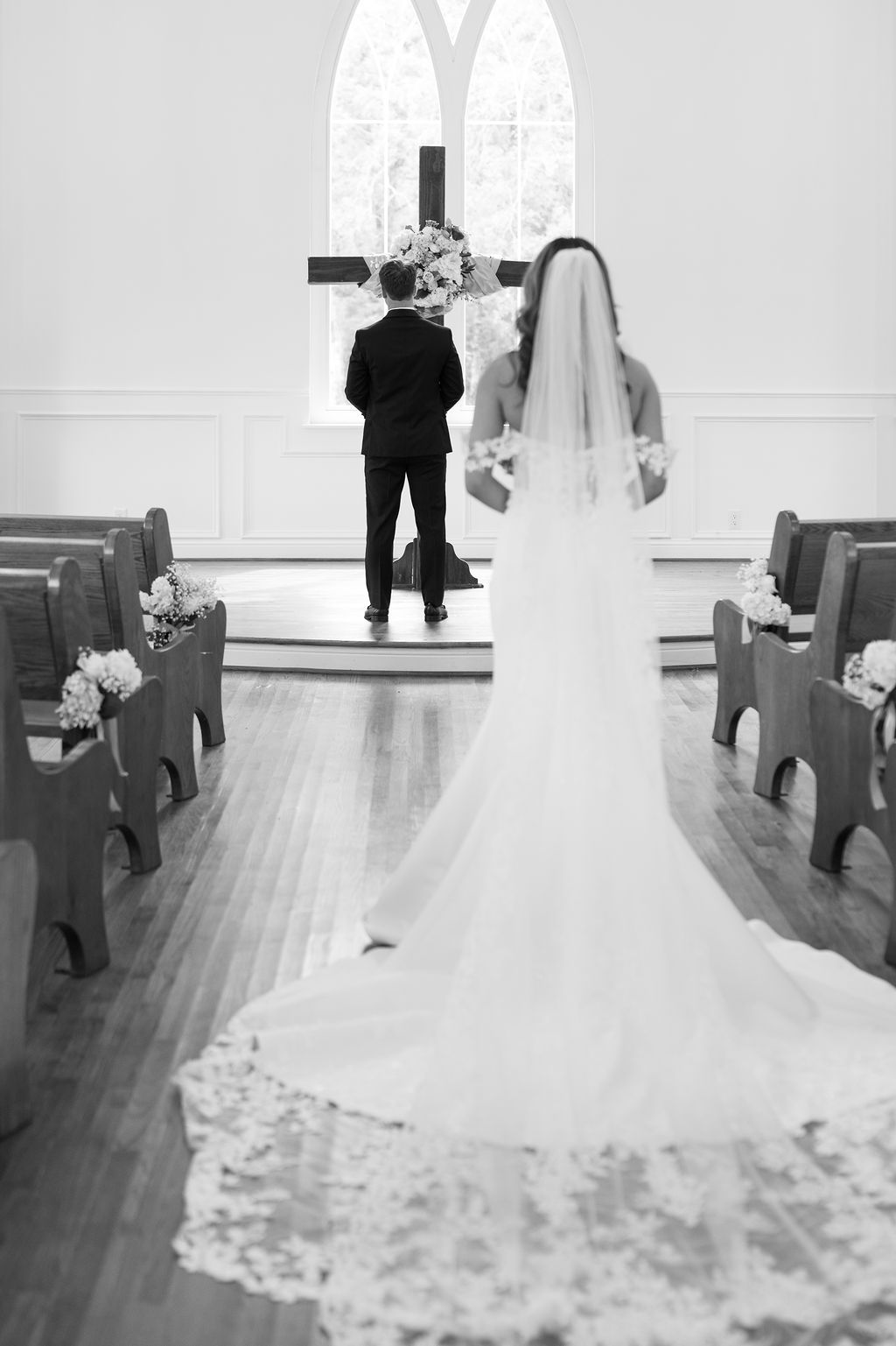 Black and white photo of bride and groom standing at church altar with cross and floral arrangements.
