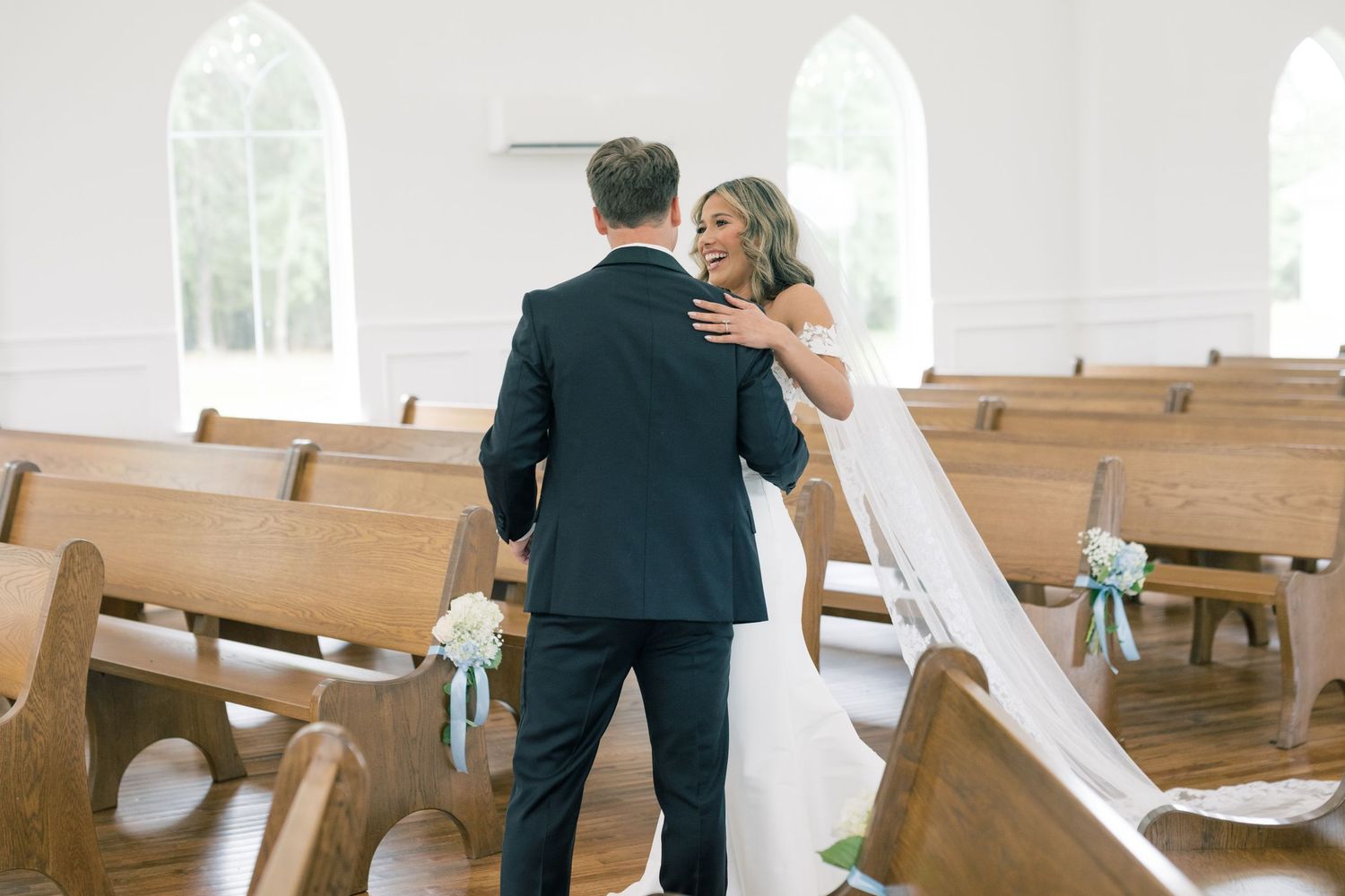 Bride and groom share first look moment inside white chapel with wooden pews and arched windows.