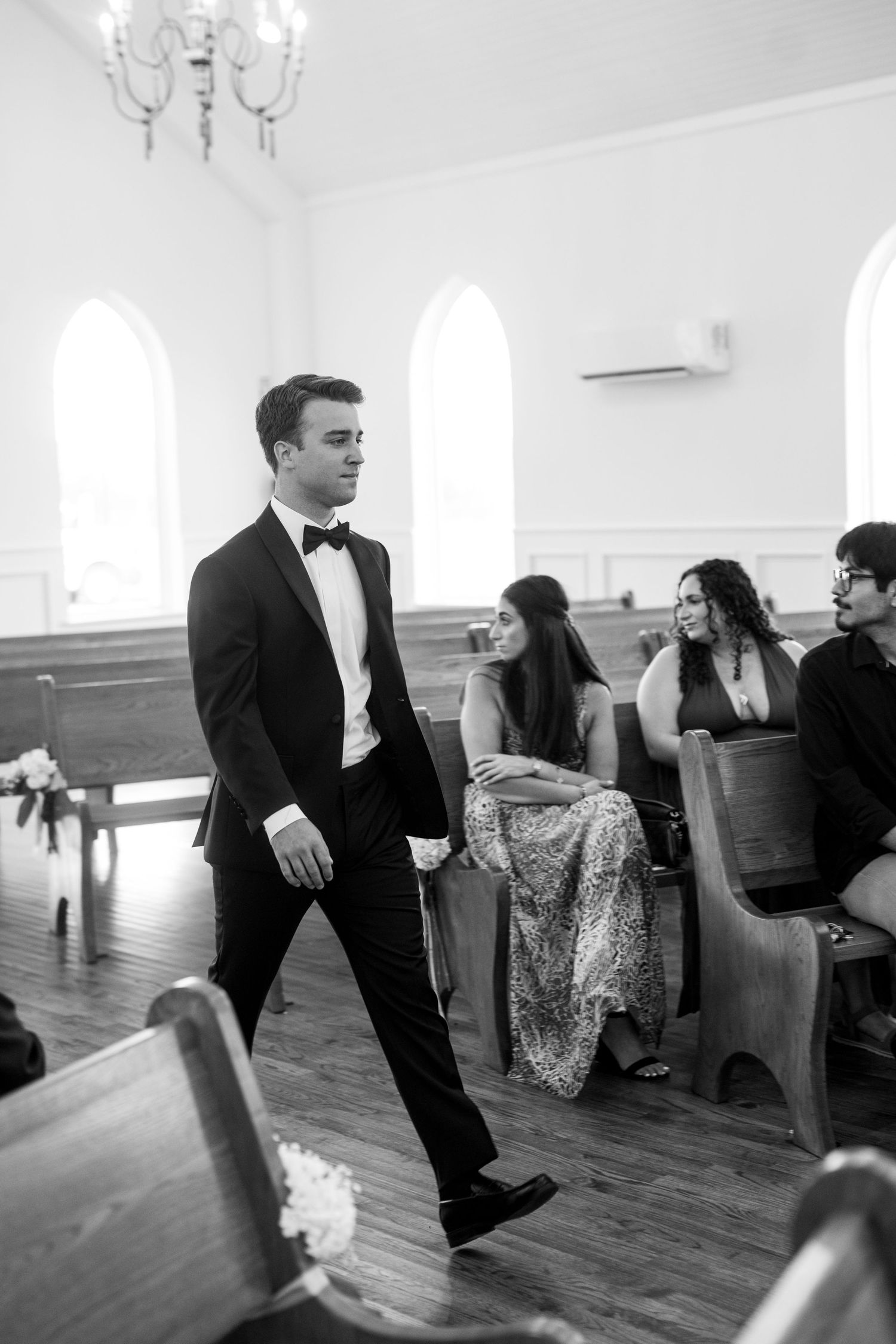 Groom in black tuxedo walking down church aisle past seated wedding guests in black and white photo.