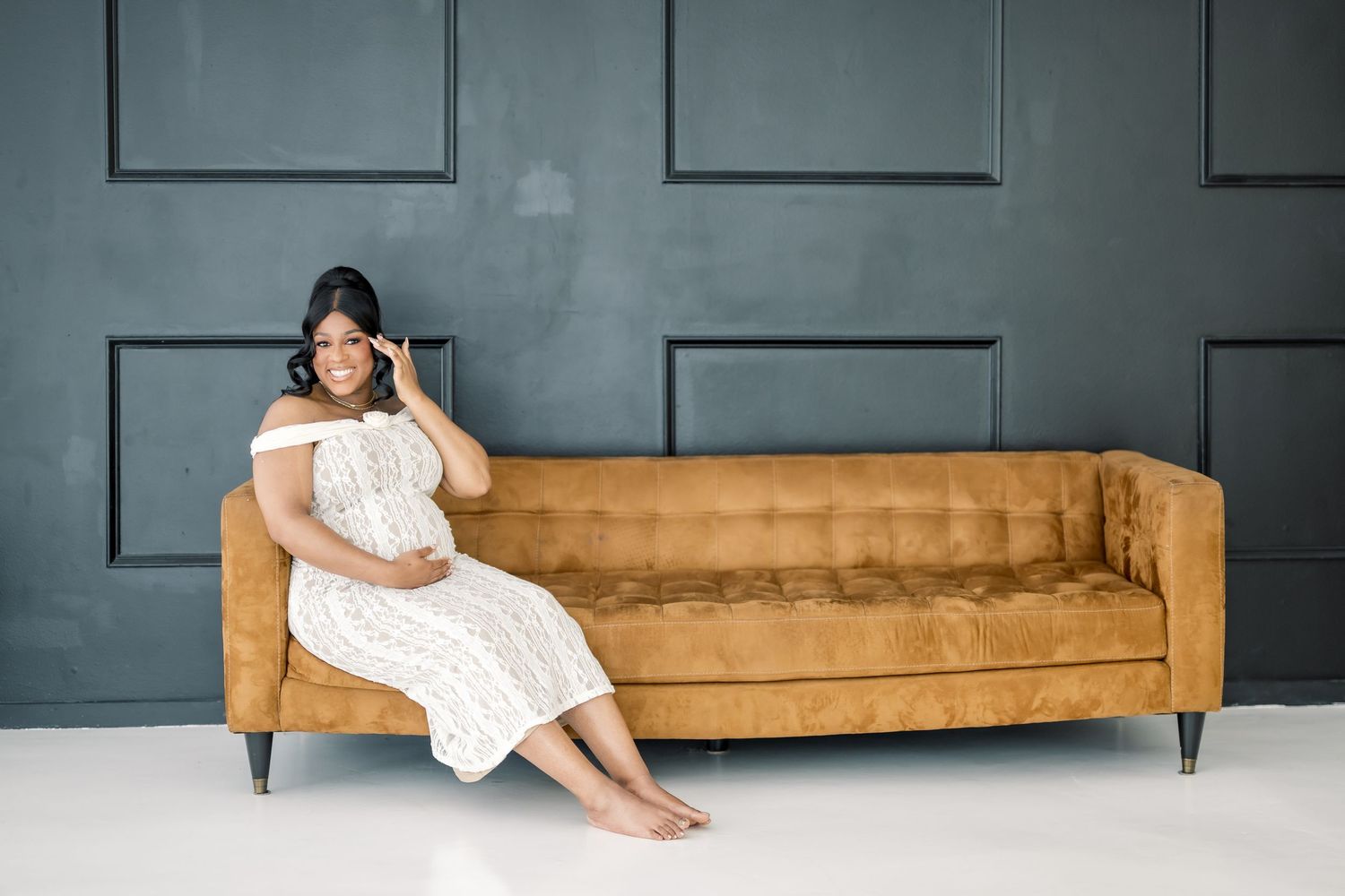 Pregnant woman in white lace dress sitting on gold velvet sofa against dark gray paneled wall.