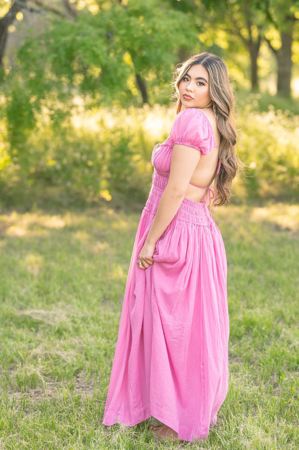 Young woman in pink maxi dress with open back poses outdoors in lush green park setting.