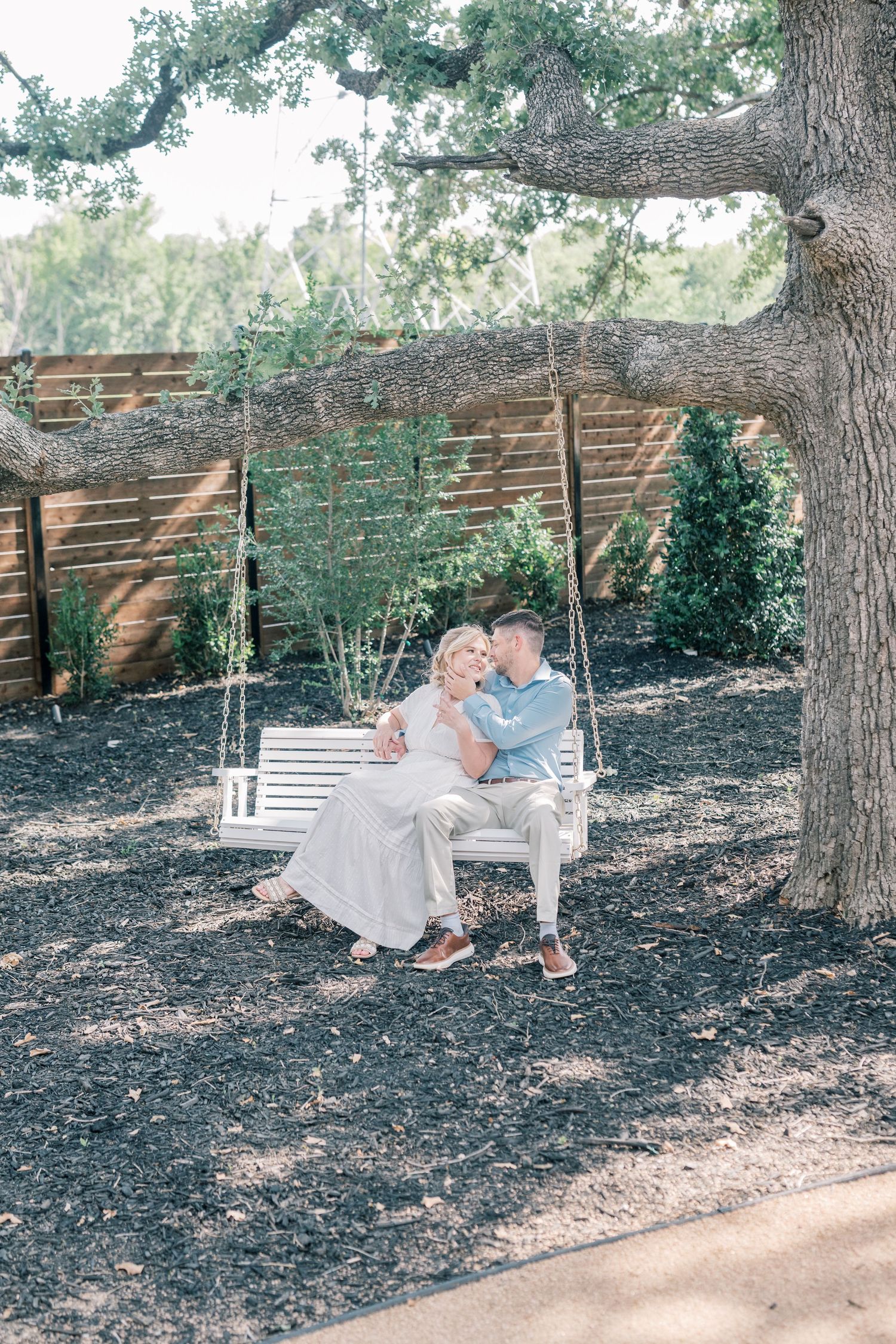 Romantic couple sitting close together on a white garden swing beneath a large shady oak tree.