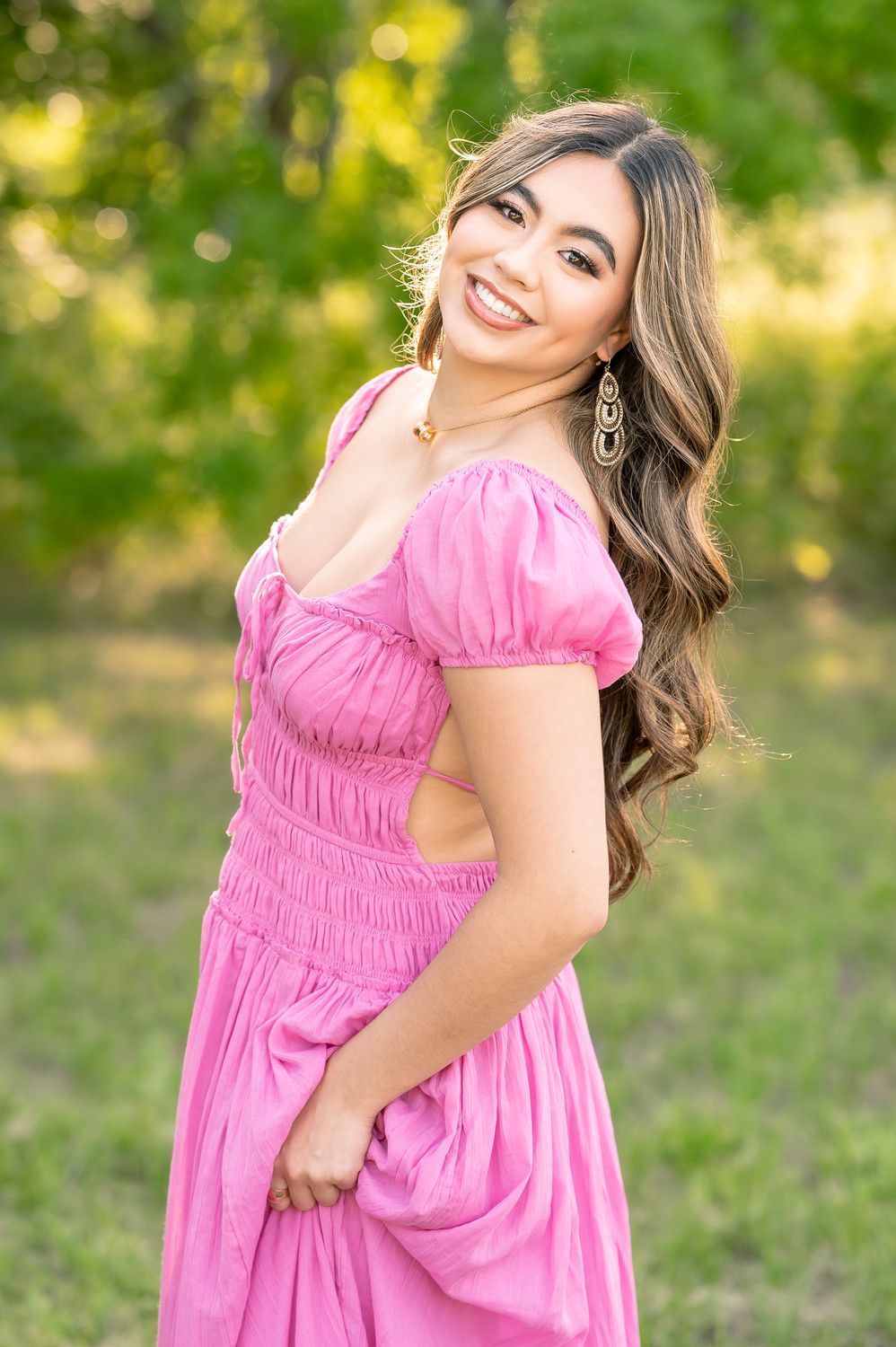 Young woman in pink off-shoulder dress smiling outdoors with green trees background. Wichita Falls & the surrounding North Texas