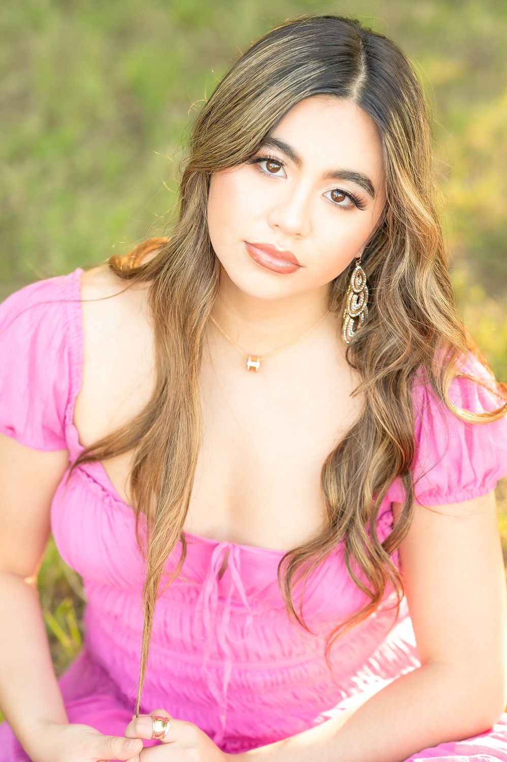 Young woman in pink dress with wavy hair, gold earrings, and necklace poses outdoors in a portrait photo.