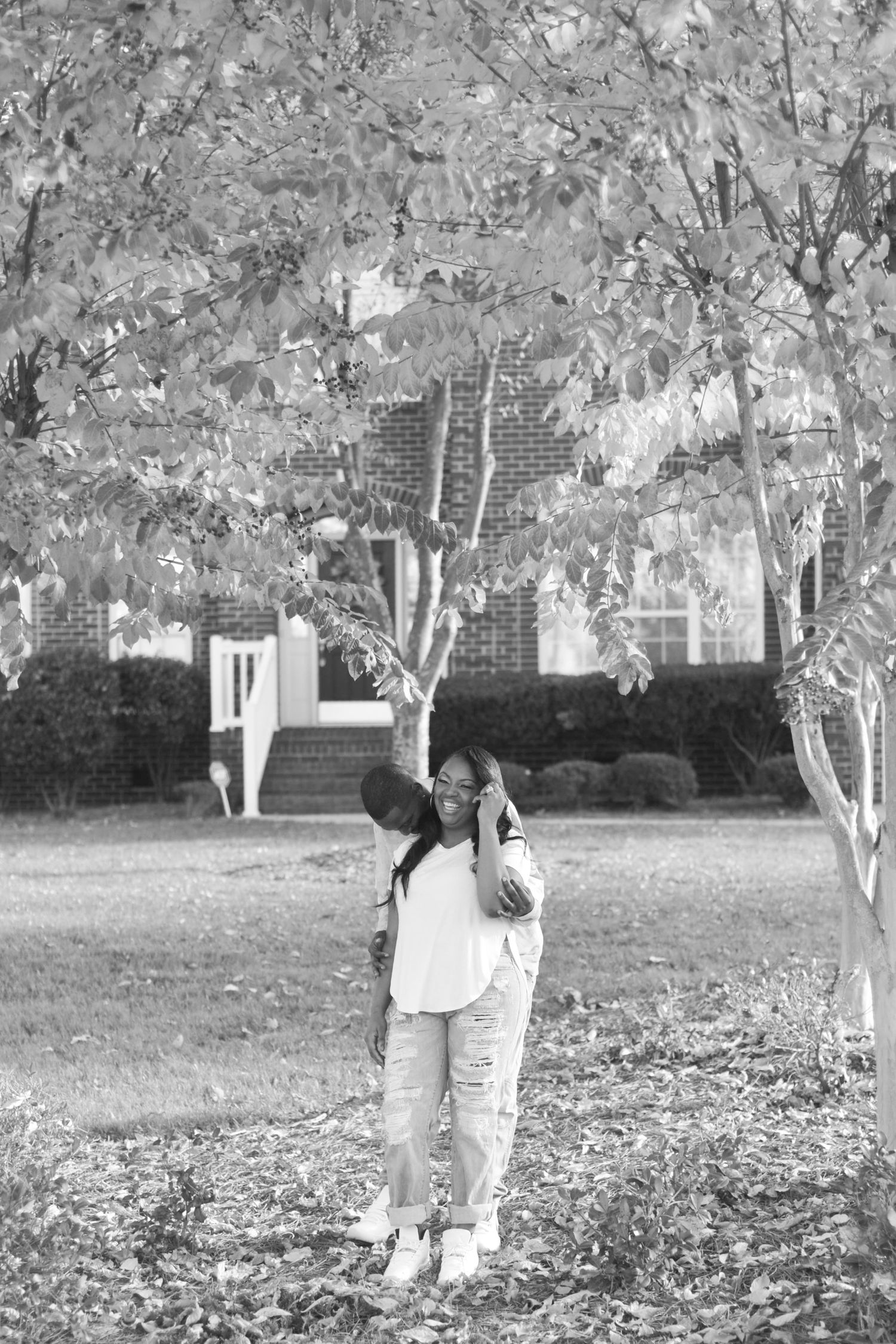 Black and white photo of a couple embracing tenderly under a leafy tree in a park.