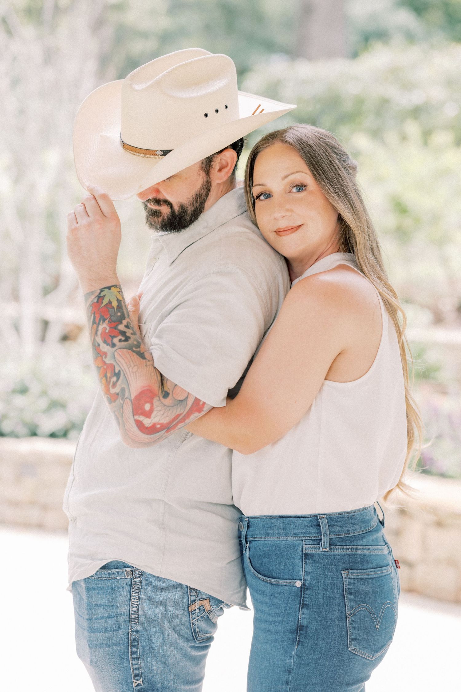 A bearded man in a cowboy hat with tattoos embraces a woman in a white tank top and jeans in an outdoor setting.