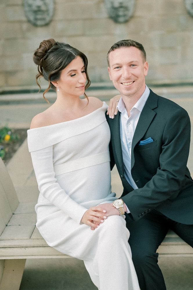 Smiling engaged couple sitting together on stone steps, woman in white off-shoulder dress and man in dark suit.