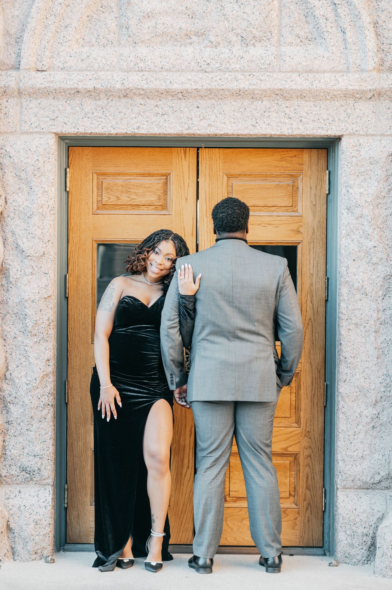 Woman in black velvet gown poses playfully as man in gray suit faces wooden doors of a stone building.