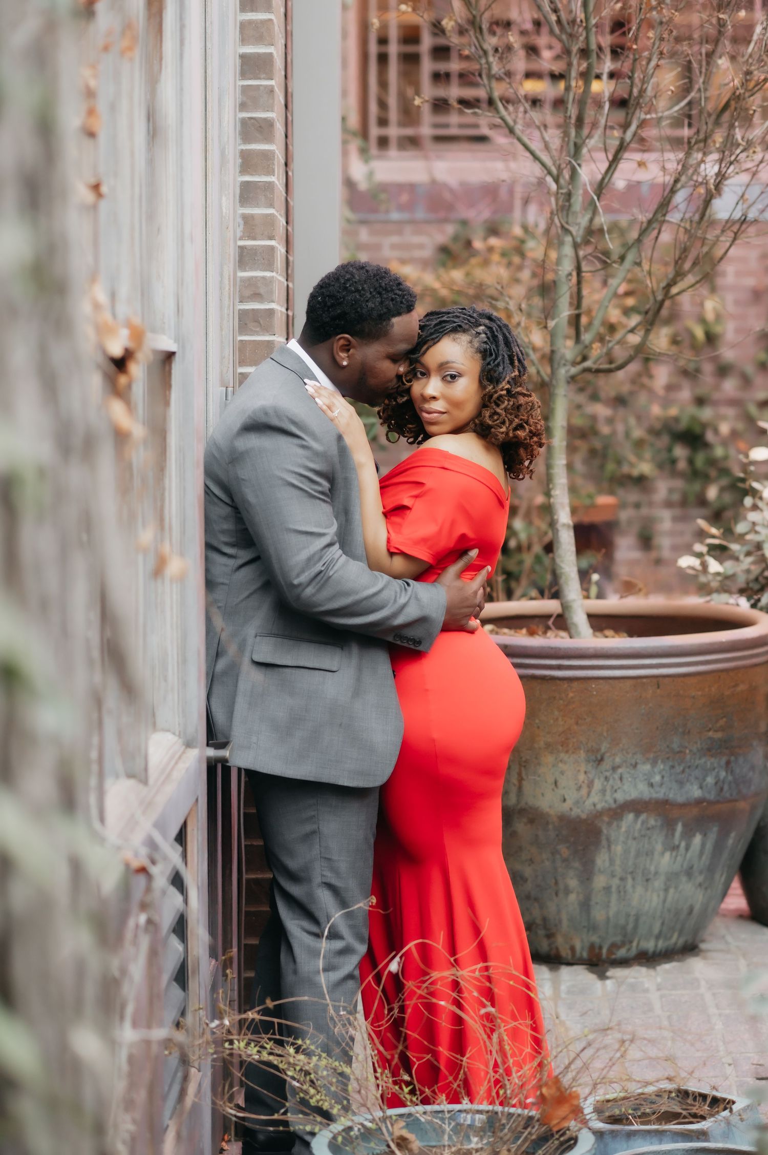Couple in red maternity gown and gray suit share tender embrace in elegant outdoor courtyard setting.