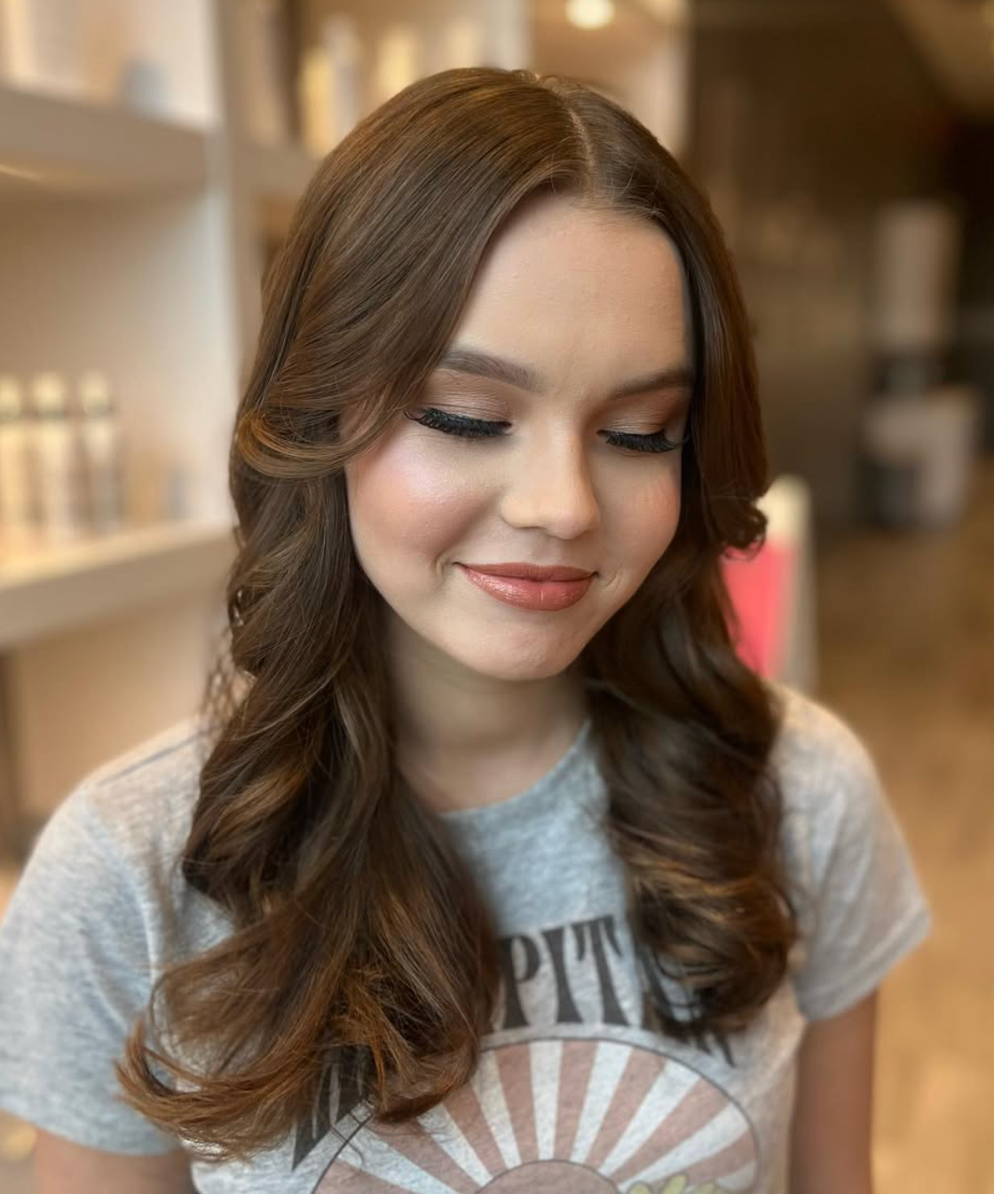 Young woman with styled brown wavy hair and natural makeup smiling softly in a salon setting.