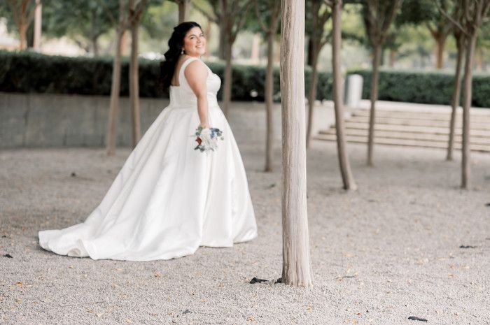 Bride in a flowing white ball gown with a long train holds a bouquet among trees on a gravel path.