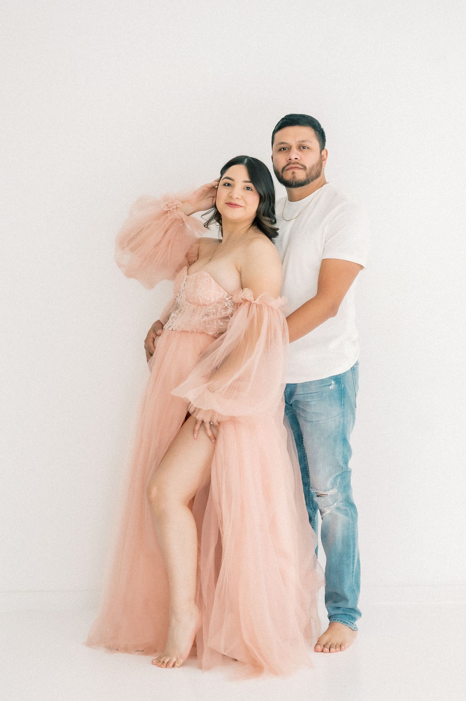 Pregnant woman in pink tulle gown poses with partner in white tee and jeans against white studio backdrop.